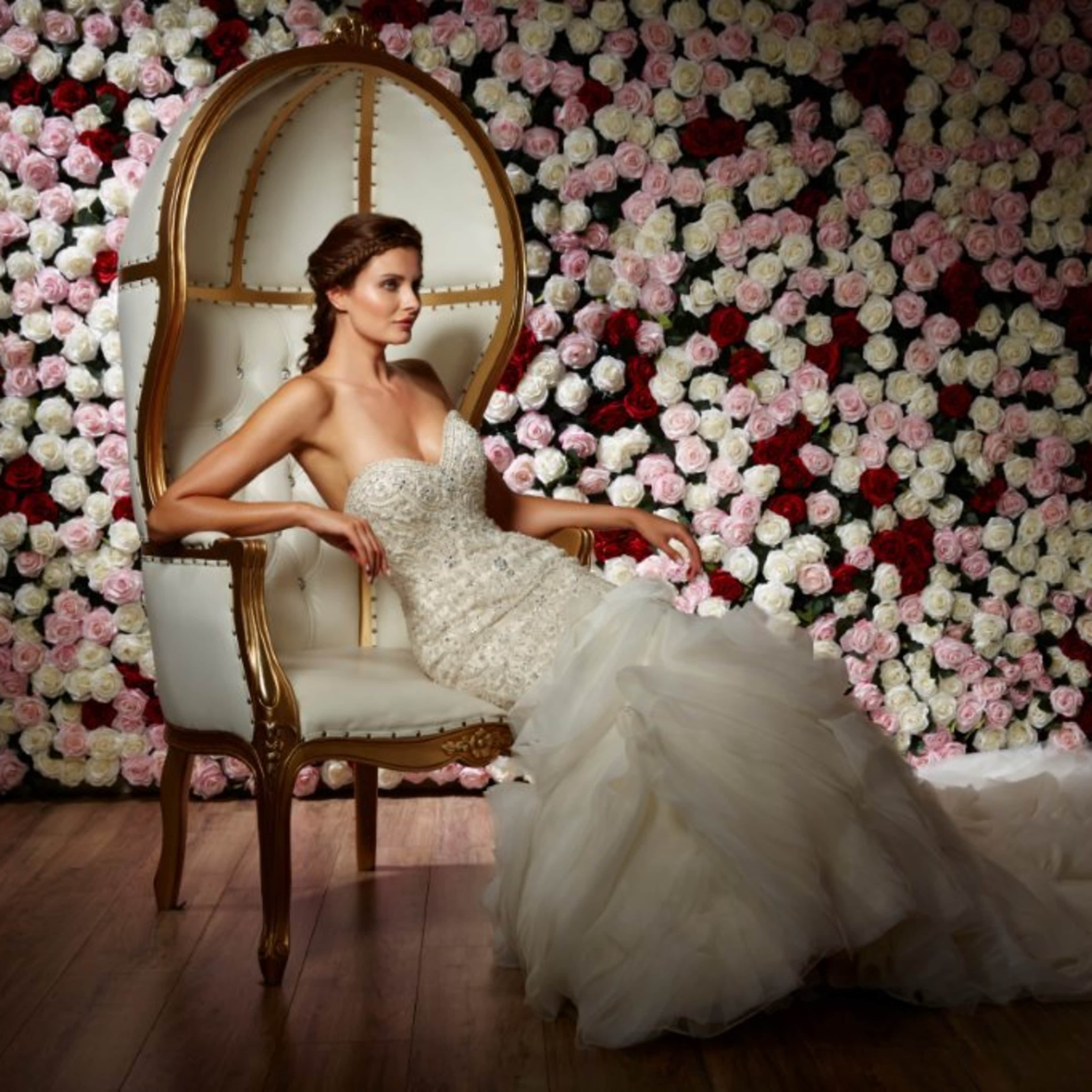 A woman in a wedding dress sits elegantly on a large chair in front of a wall covered with rose flowers.