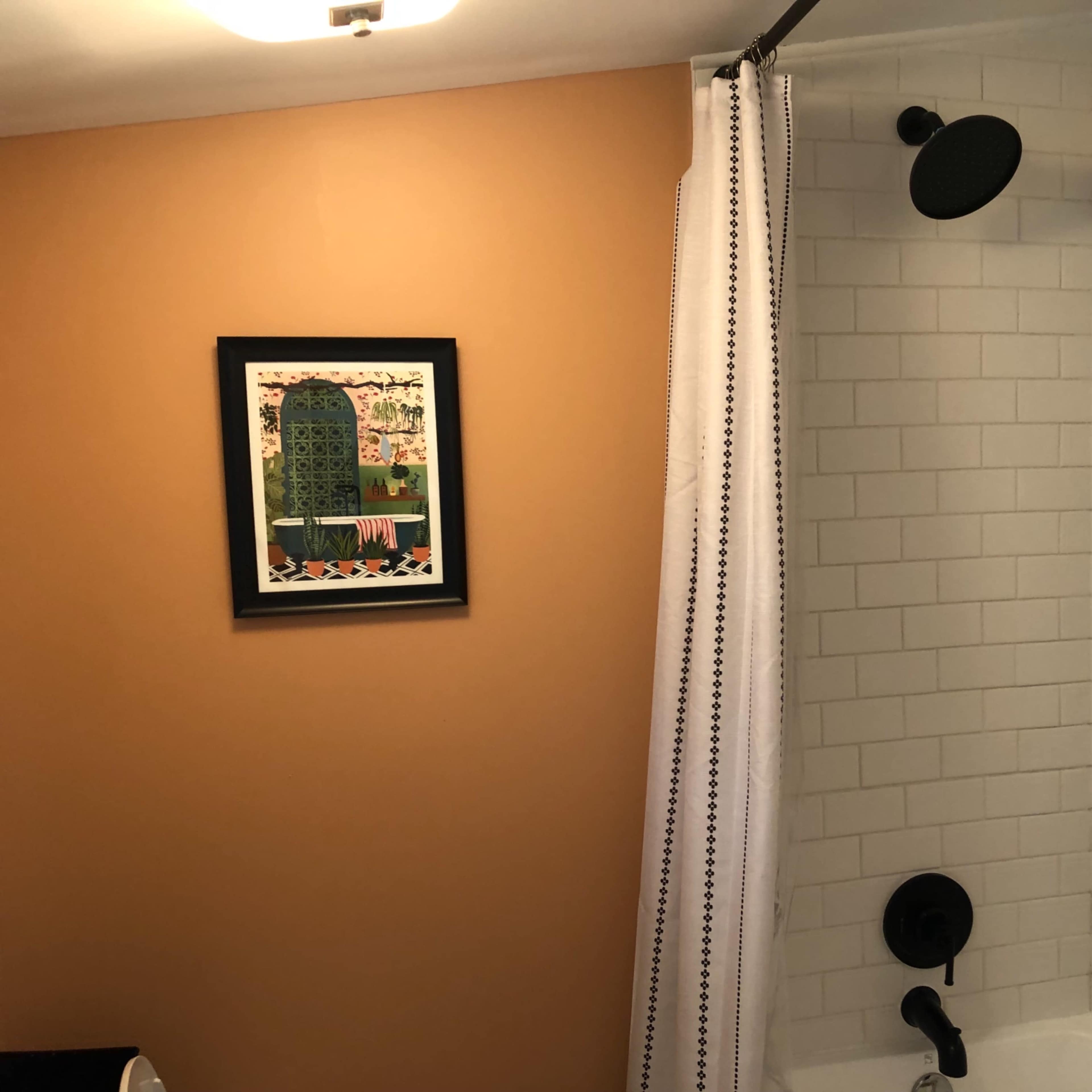 A bathroom area featuring a shower with a showerhead, white tiled walls, a curtain, and a colorful framed artwork on the orange wall.
