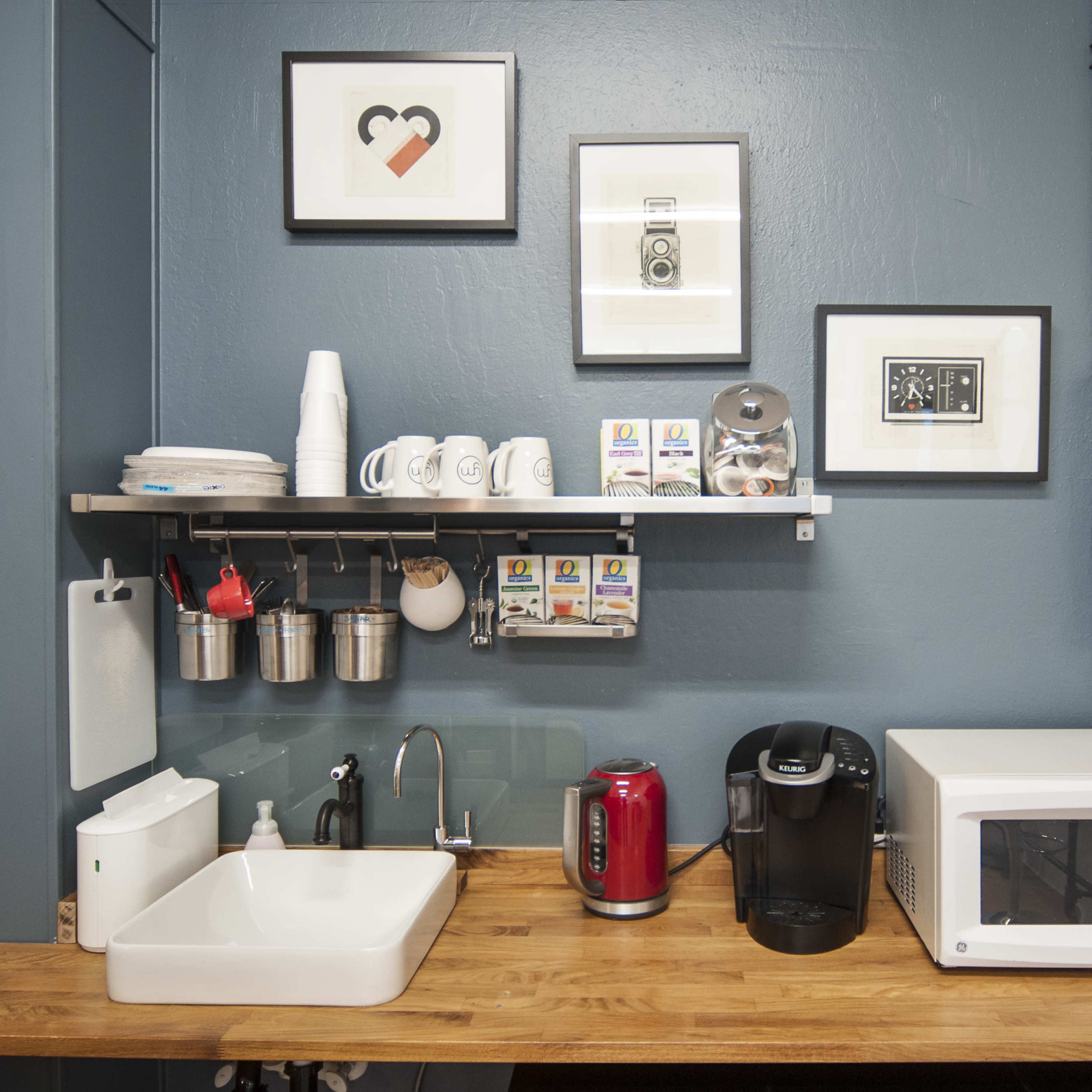 A kitchen area featuring a wooden counter with a sink, a microwave, a coffee maker, and several mugs on a shelf, against a blue wall adorned with framed artwork.