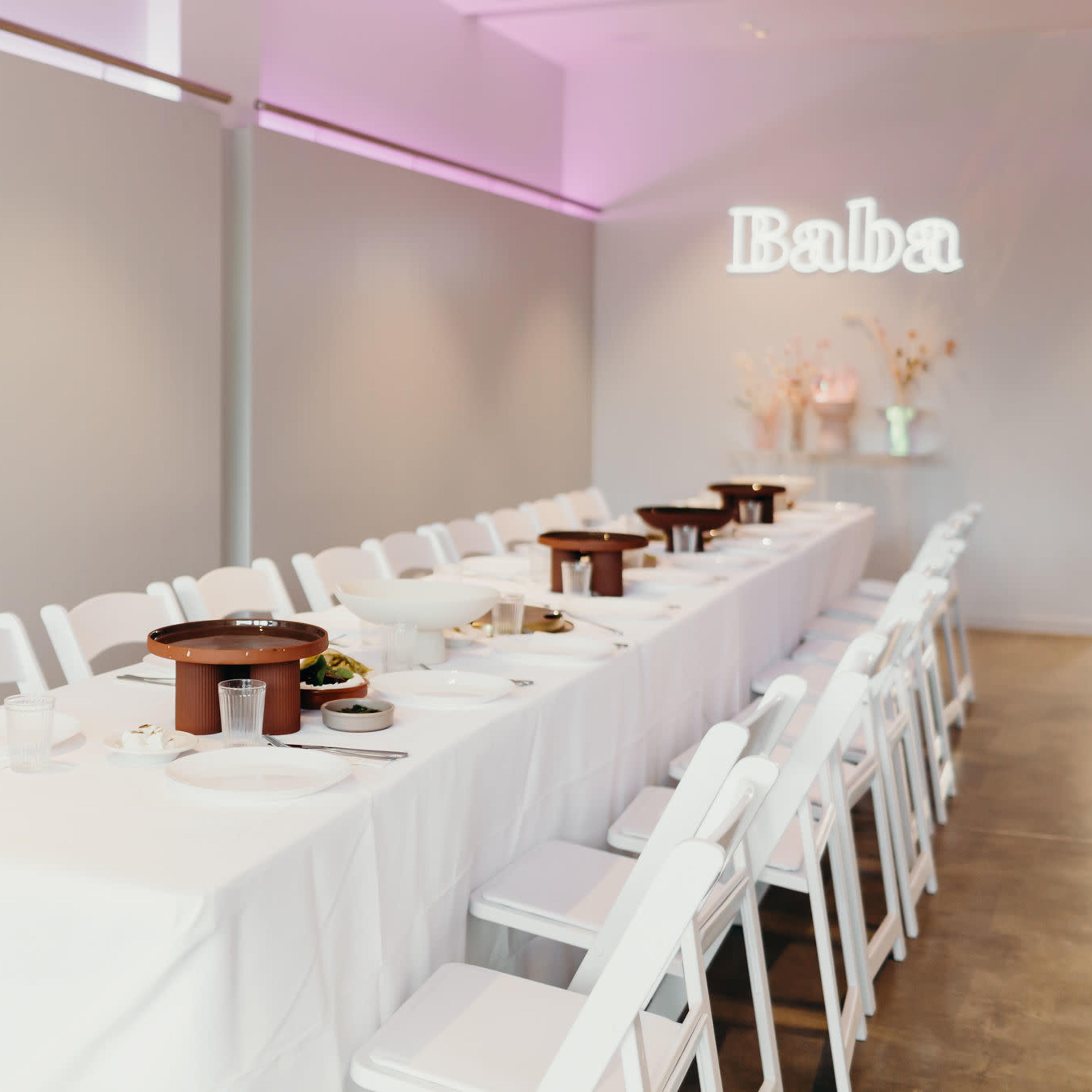 A long dining table with white tablecloth, set with plates and utensils, is positioned in a well-lit, minimalist room featuring a decorative wall and a neon sign that reads "Baba."