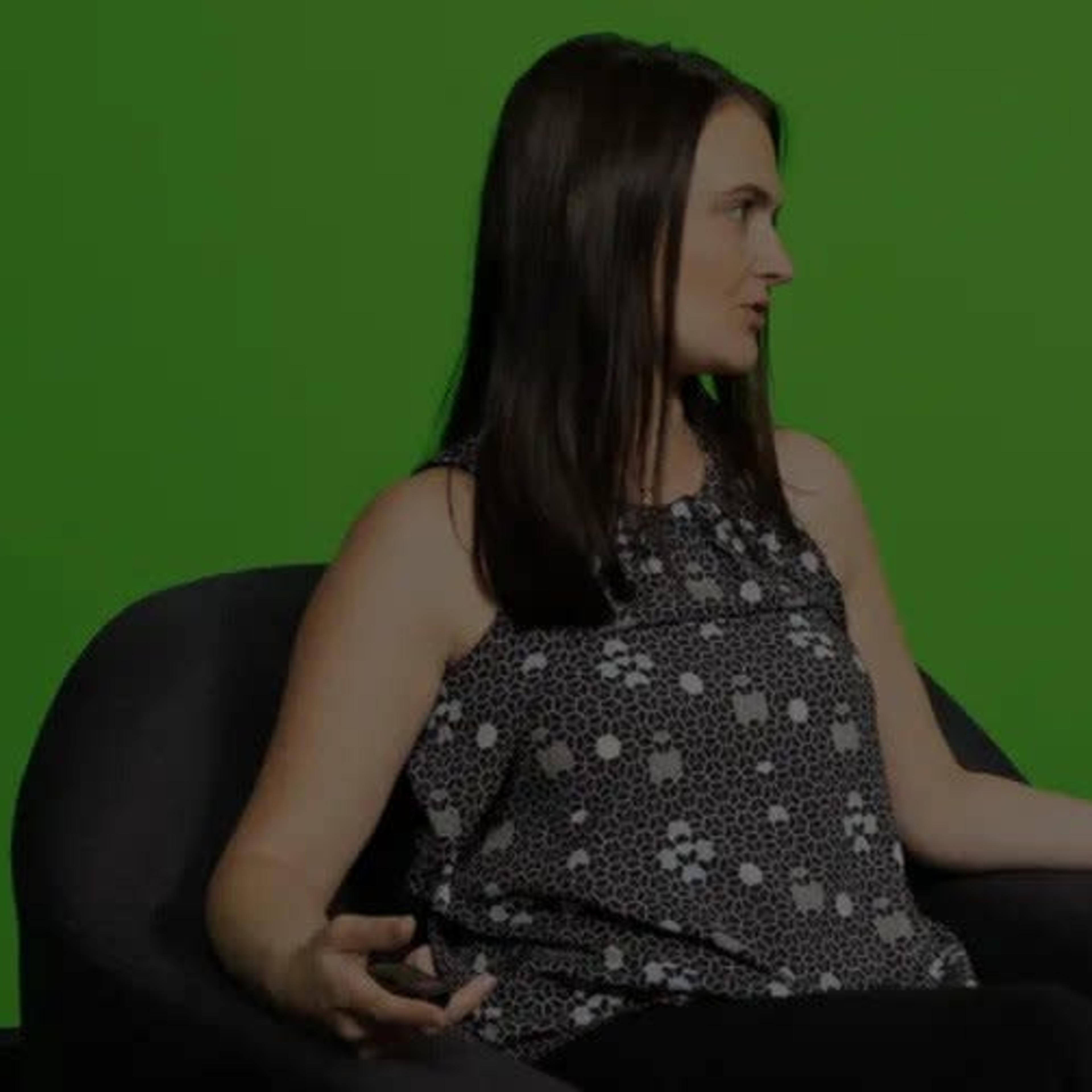 A woman sits on a gray chair in front of a green screen, gesturing with one hand.