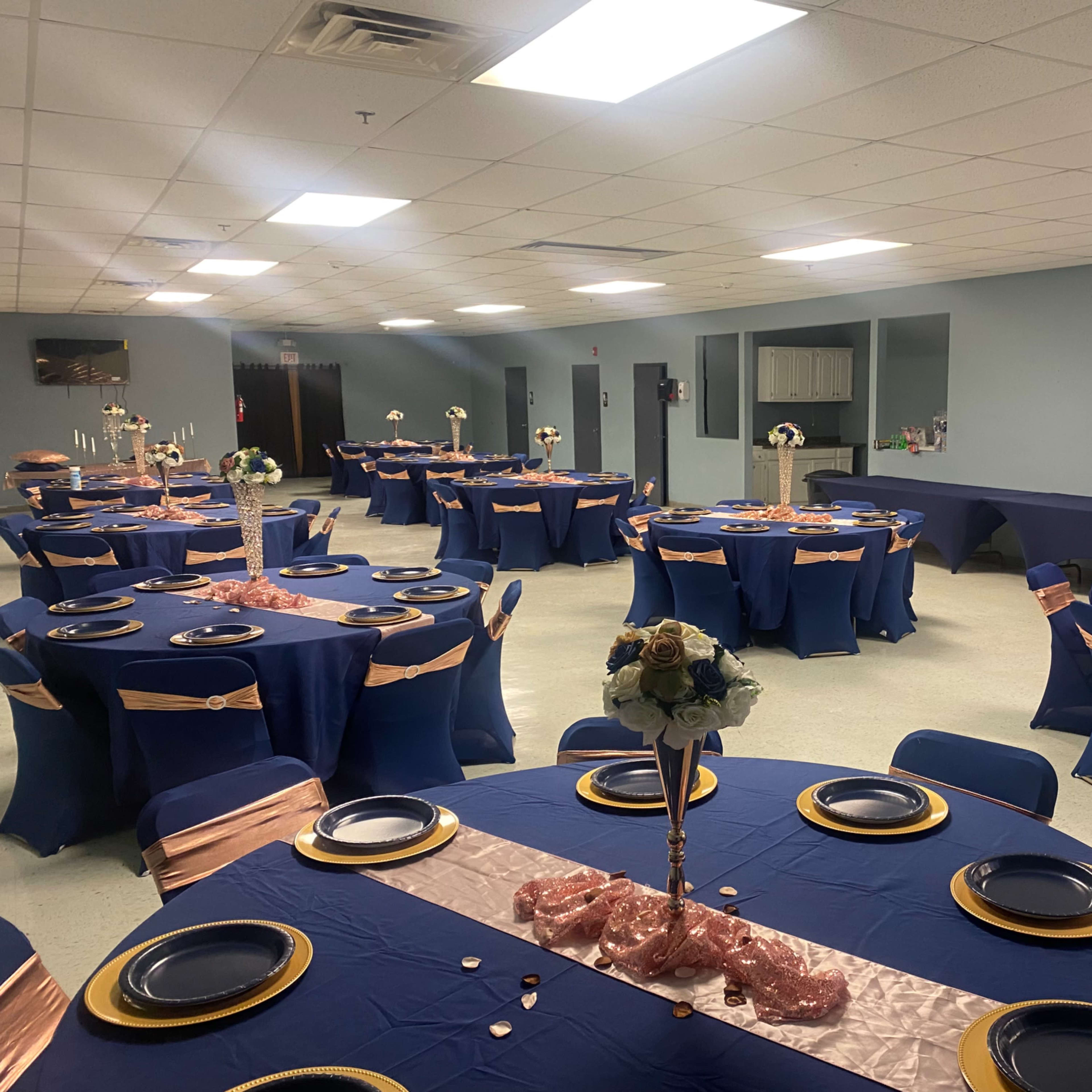 A banquet hall set with several round tables covered in navy blue tablecloths and adorned with pink sashes, each featuring gold plates and floral centerpieces.