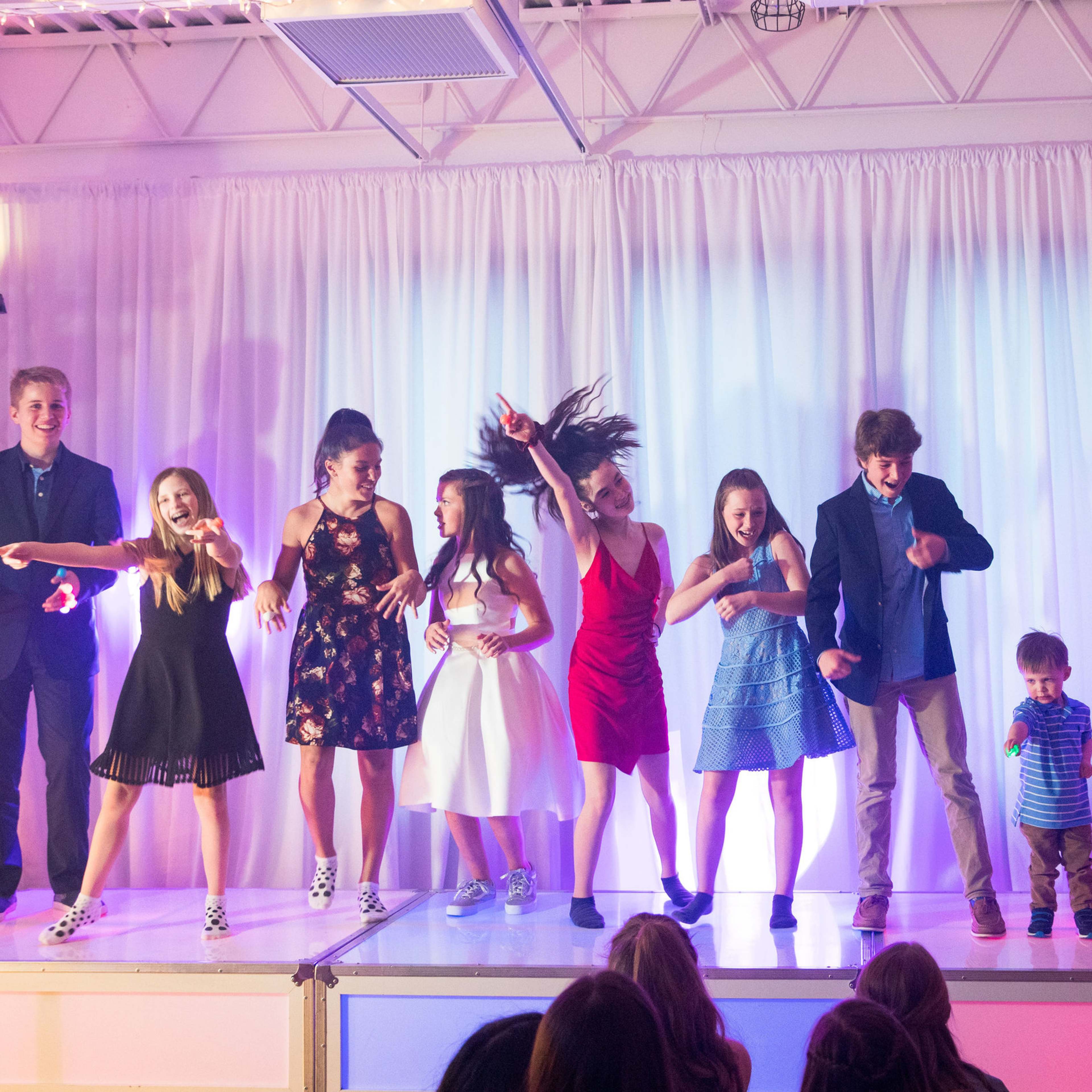 A group of children and teenagers are performing on a brightly lit stage with colorful background lighting.