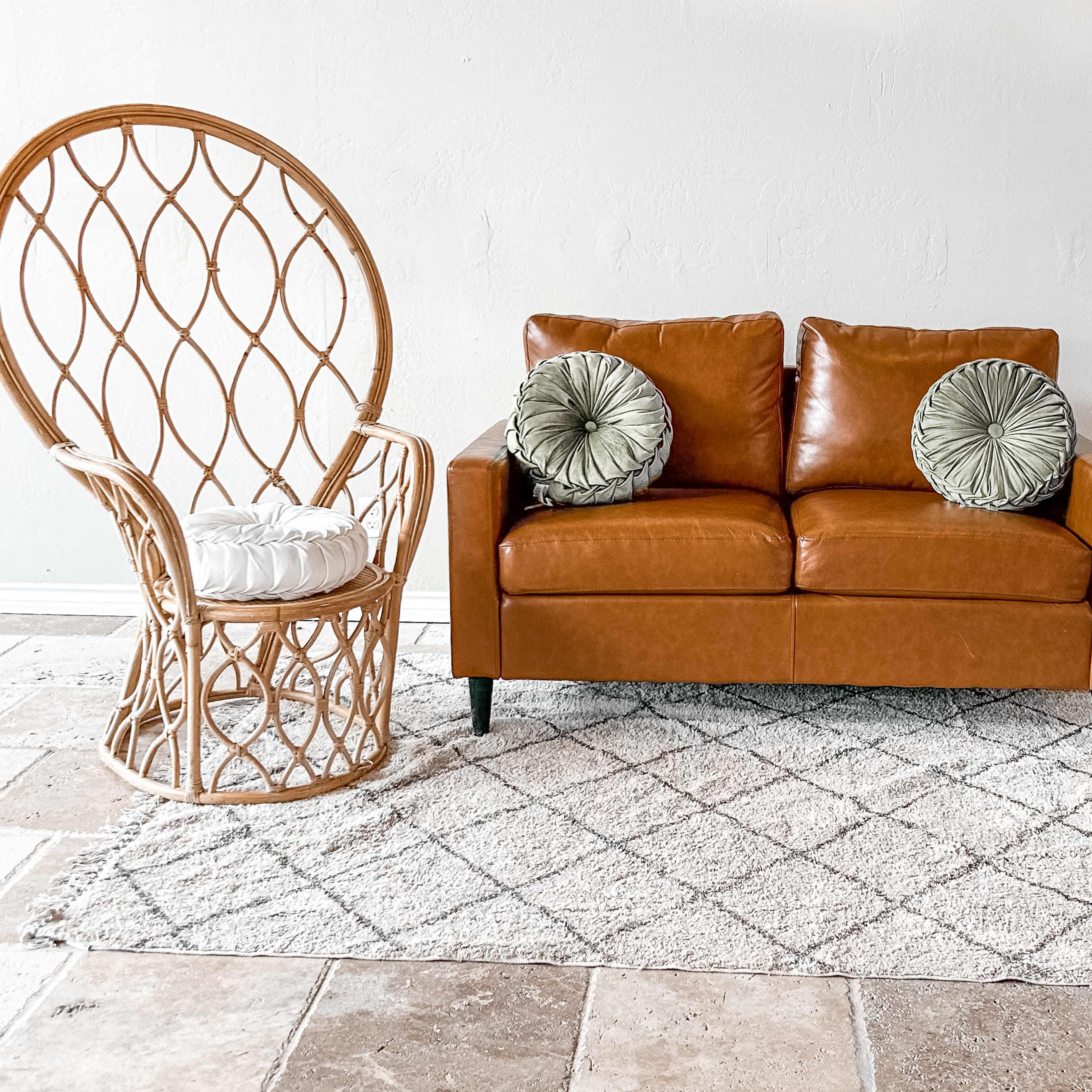 A brown leather sofa with two cushions, a rattan chair, a small side table, and a potted plant, all arranged on a patterned rug in a neutral-colored room.