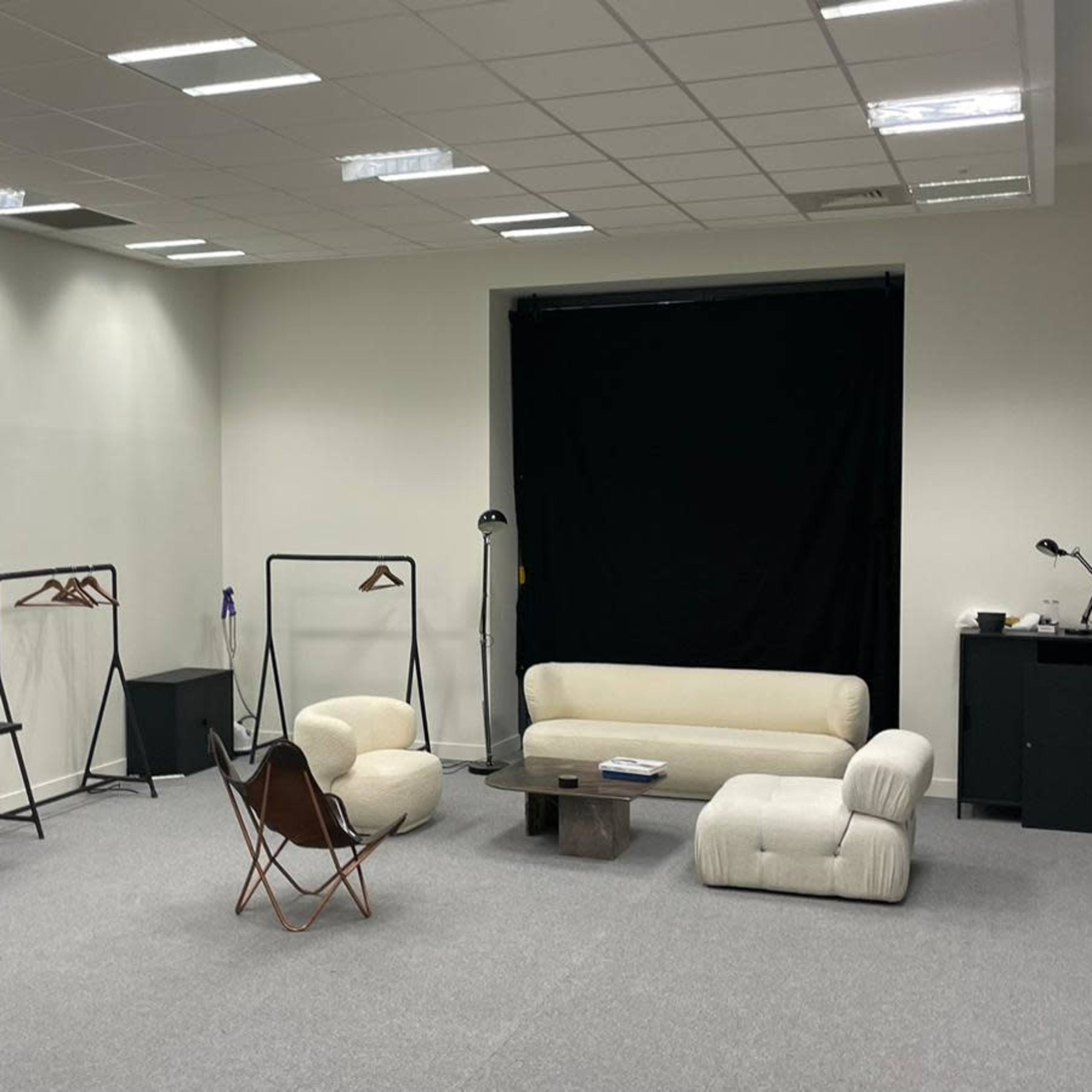 A well-lit studio space featuring a beige sofa, a small table, and various studio equipment, including light stands and backdrop supports, arranged around the area.