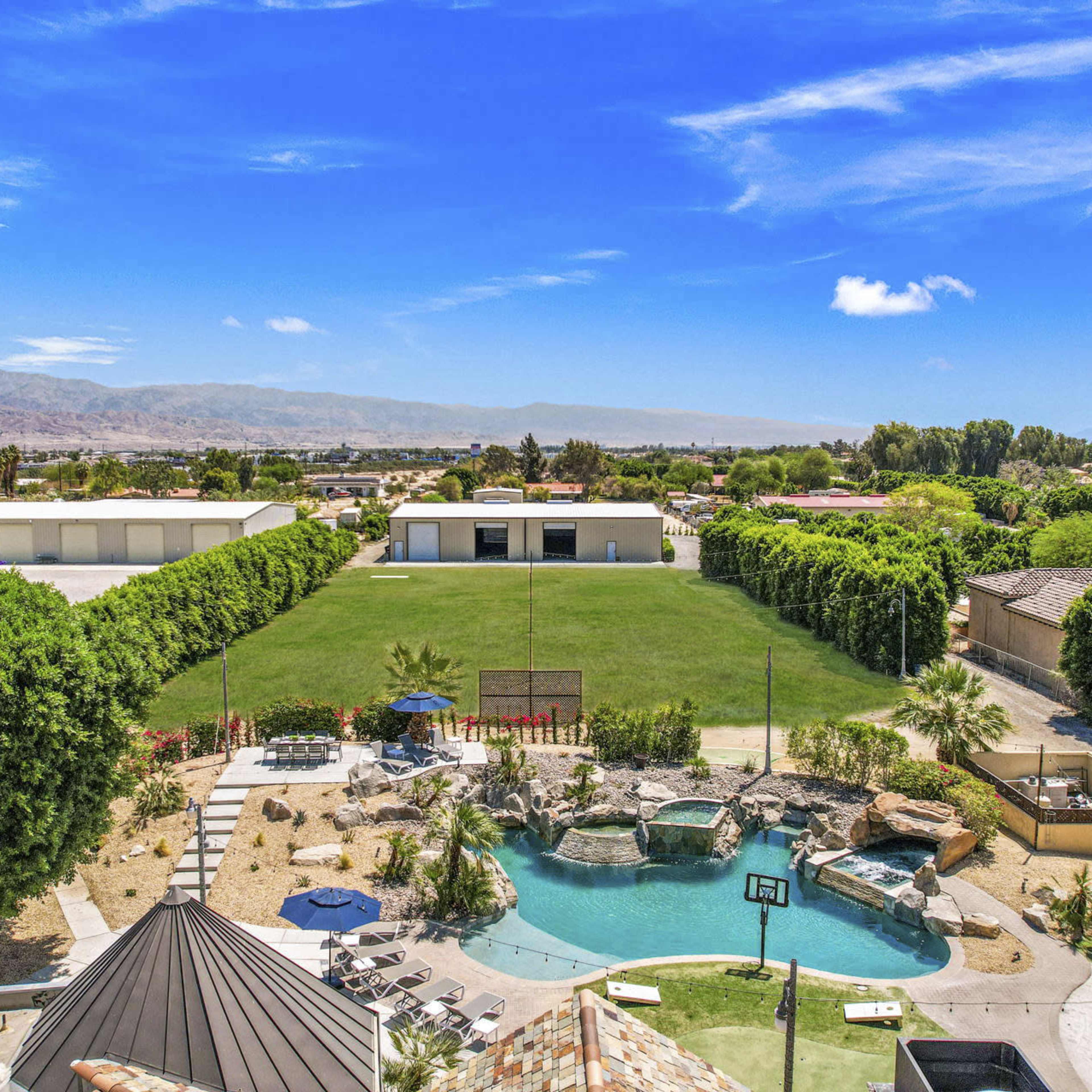 A spacious outdoor area featuring a large green lawn, several palm trees, a swimming pool with a rock formation, and multiple buildings in the background against a clear blue sky.