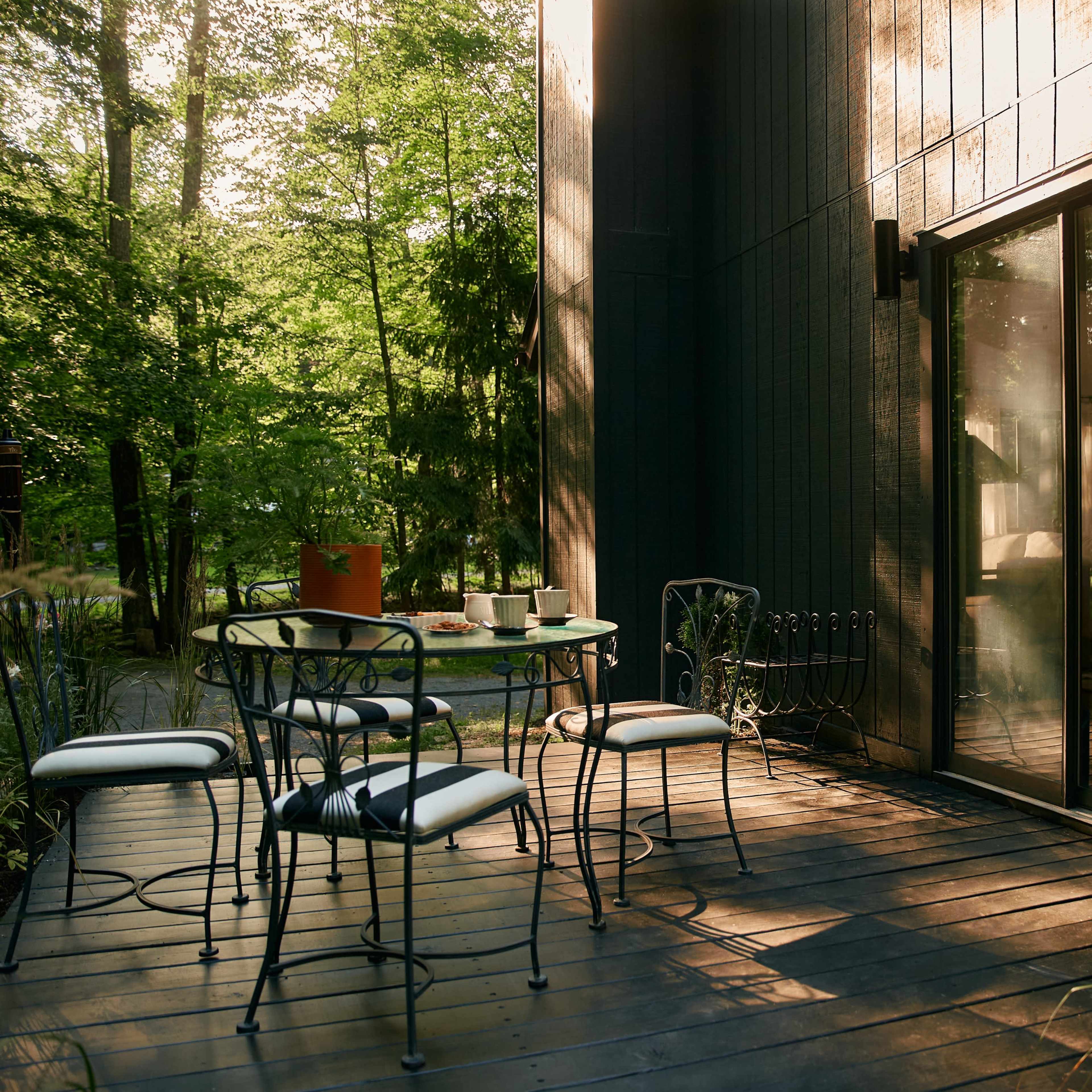 A set of metal chairs and a round table are arranged on a wooden deck beside a glass door, surrounded by greenery in soft afternoon light.