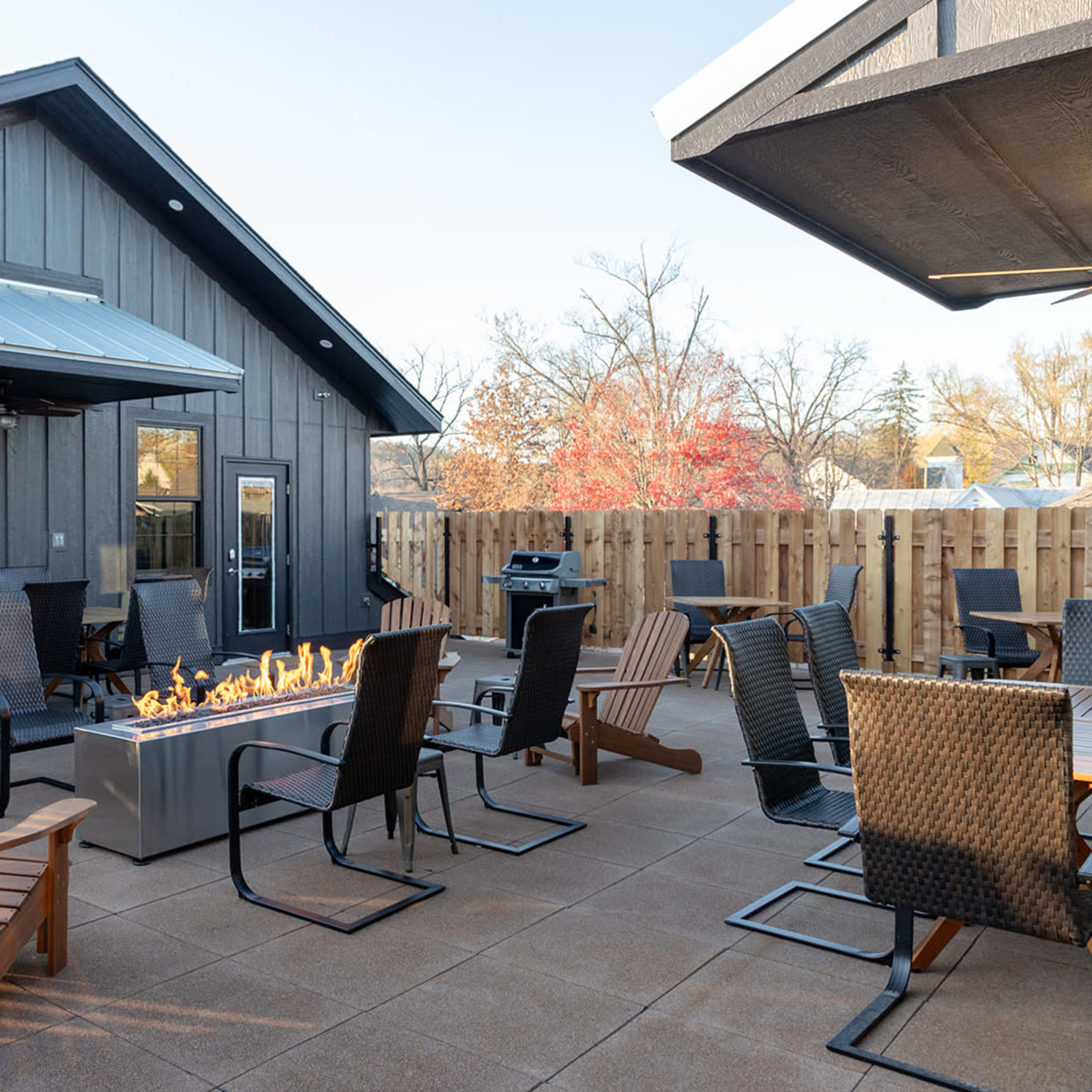 A patio area with several outdoor chairs arranged around a central fire pit, adjacent to a wooden fence and a building in the background.