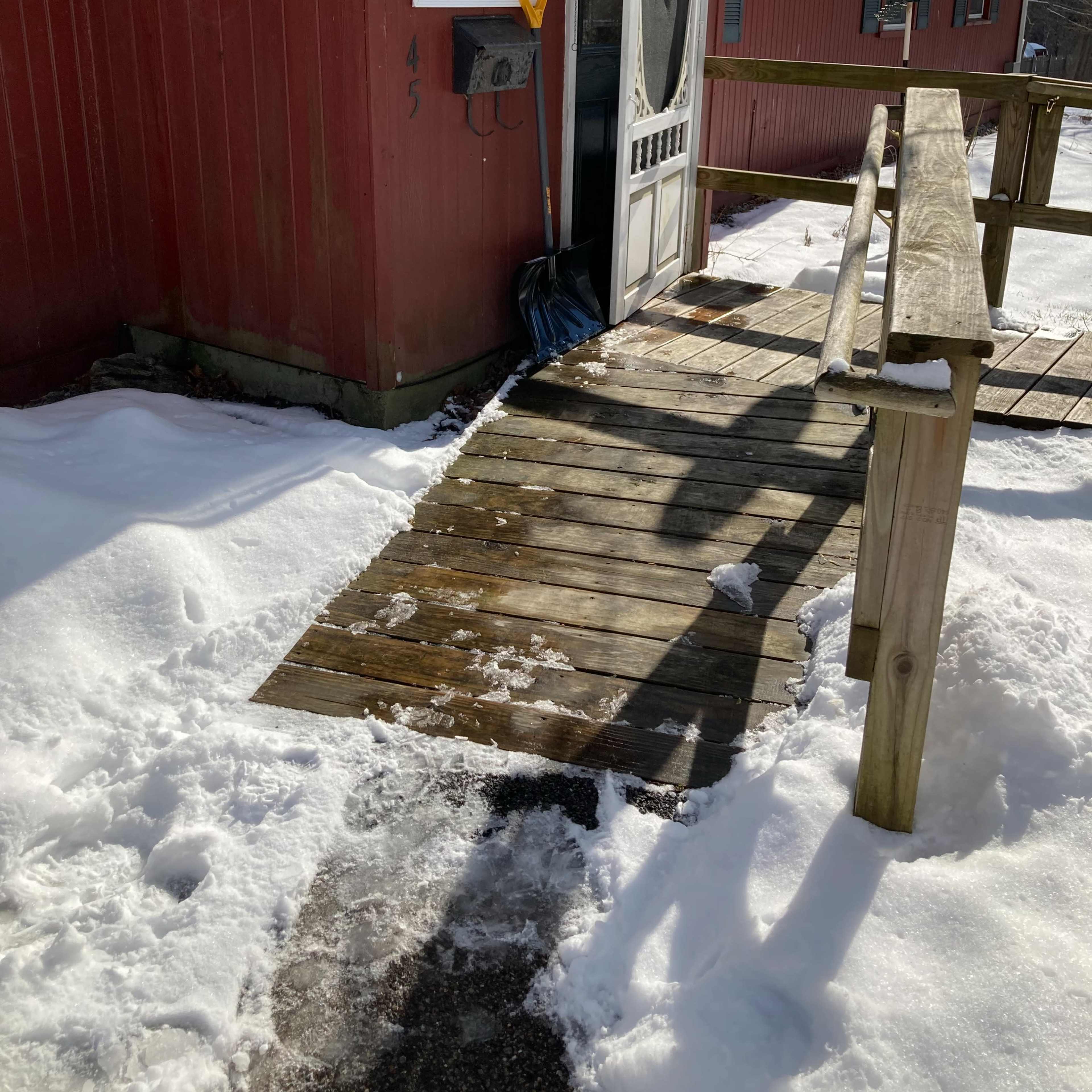 A wooden walkway leads to a red house with a storm door, surrounded by snow.