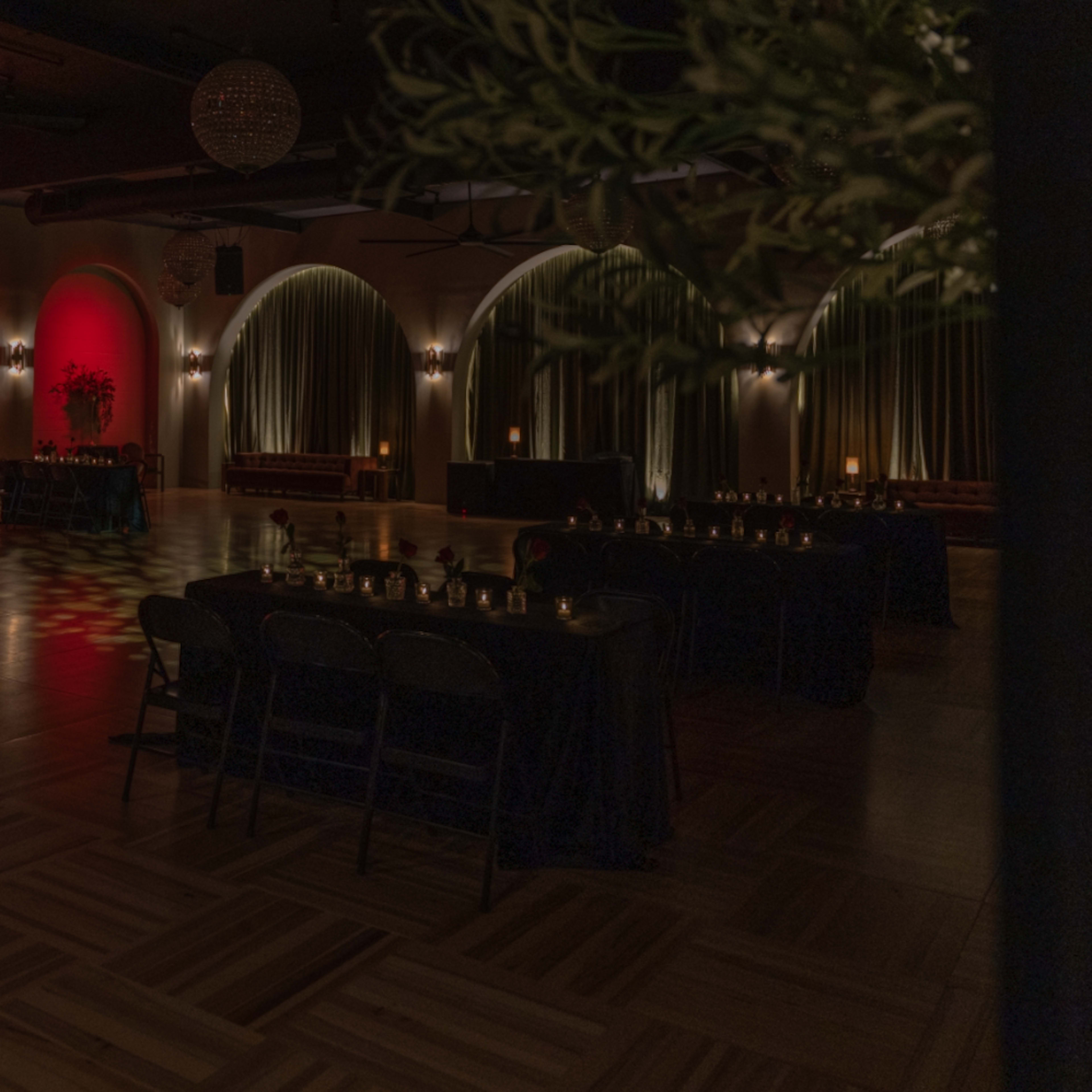 A dimly lit event space with red lighting, arched windows, and tables set for an occasion.