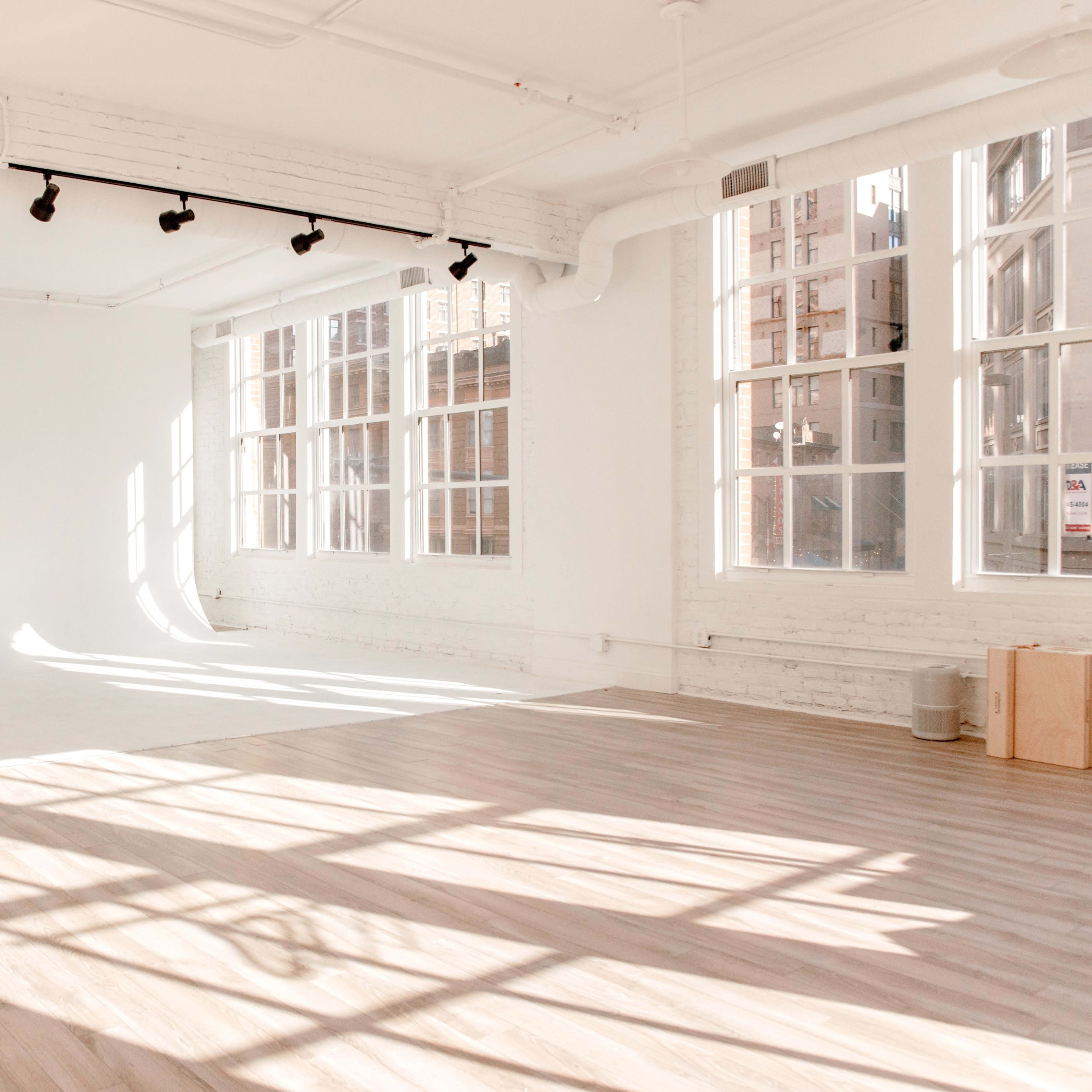 A spacious, empty room with large windows, white walls, and wooden flooring, illuminated by natural light.