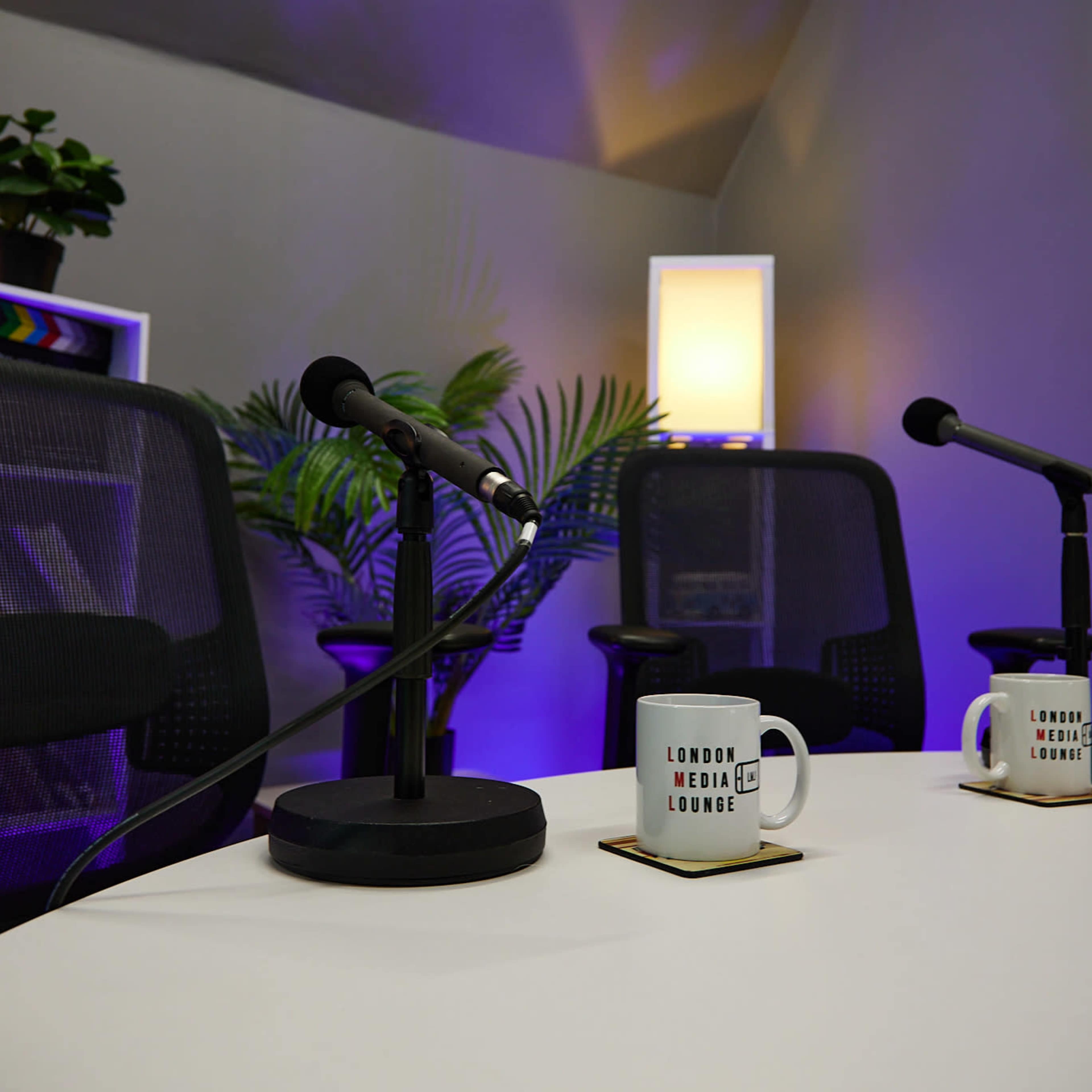 A podcasting setup with two chairs, two microphones, and mugs on a white table in a room with purple lighting.