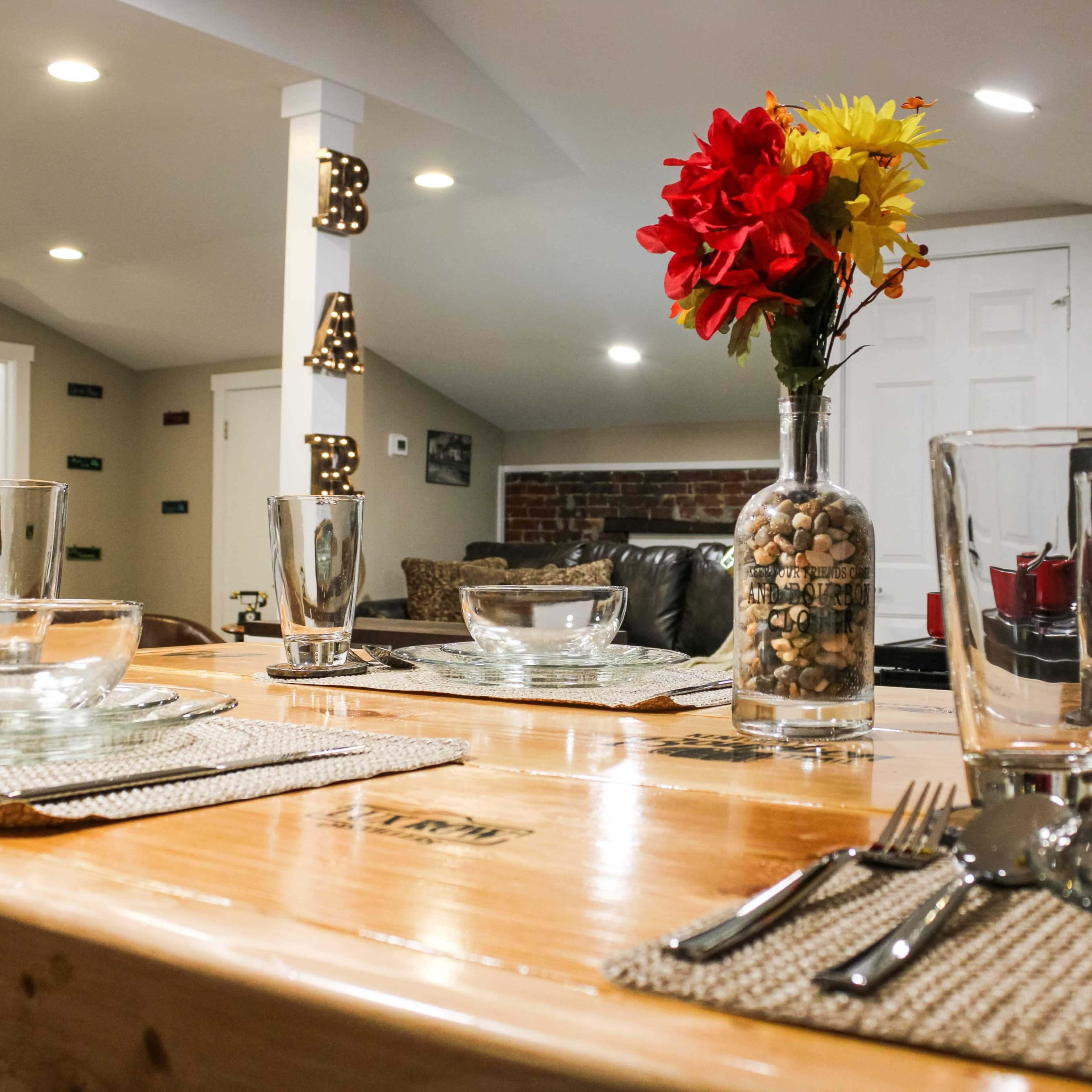 A dining table is set with glassware and utensils, featuring a vase of flowers and a rustic wooden surface, against a backdrop of a cozy living area.
