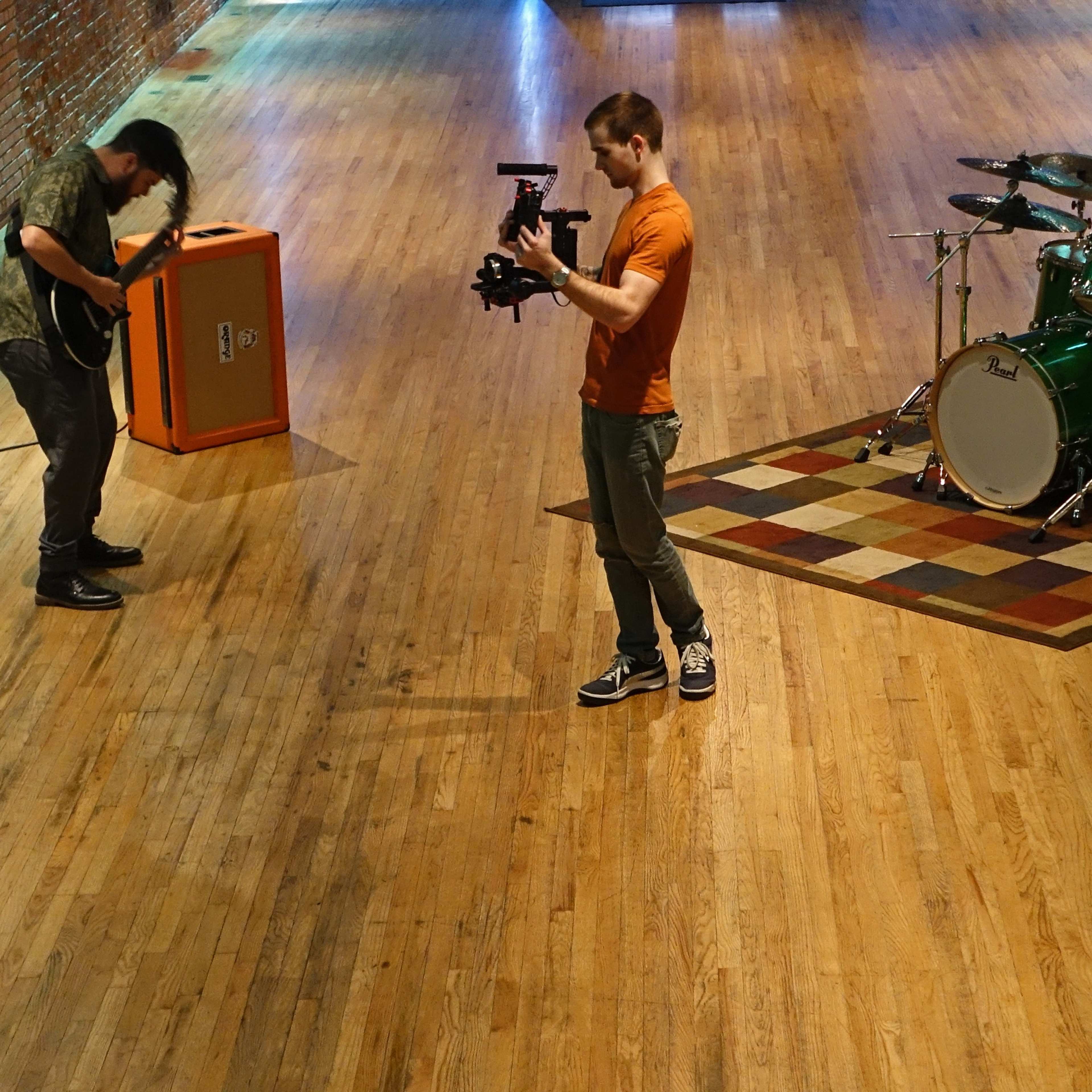 A band performs in a brick-walled studio with a female vocalist and musicians playing instruments while a cameraman records the scene.