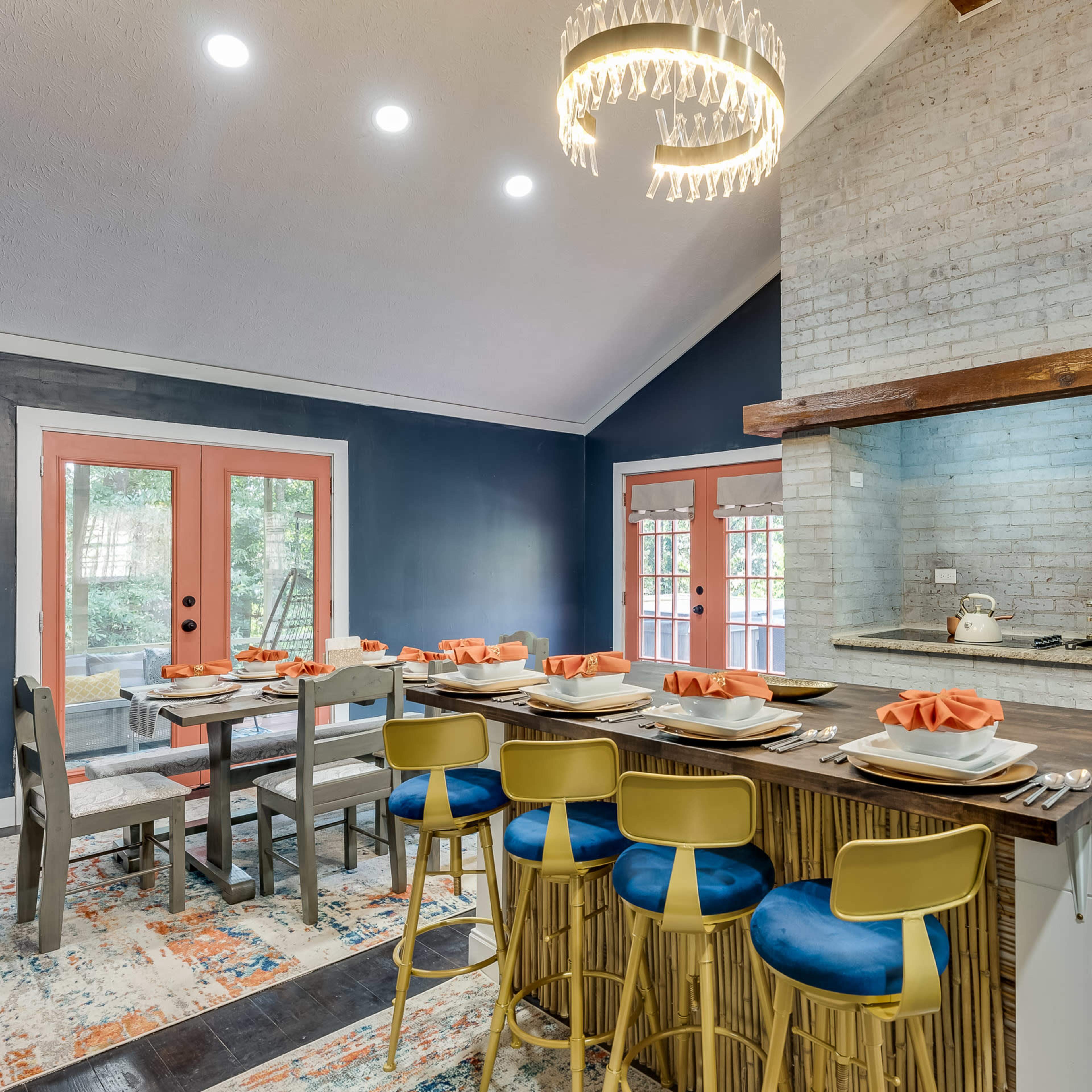 A modern kitchen features a large dining table with orange napkins and a countertop bar with blue stools, set against a backdrop of exposed brick and dark walls.