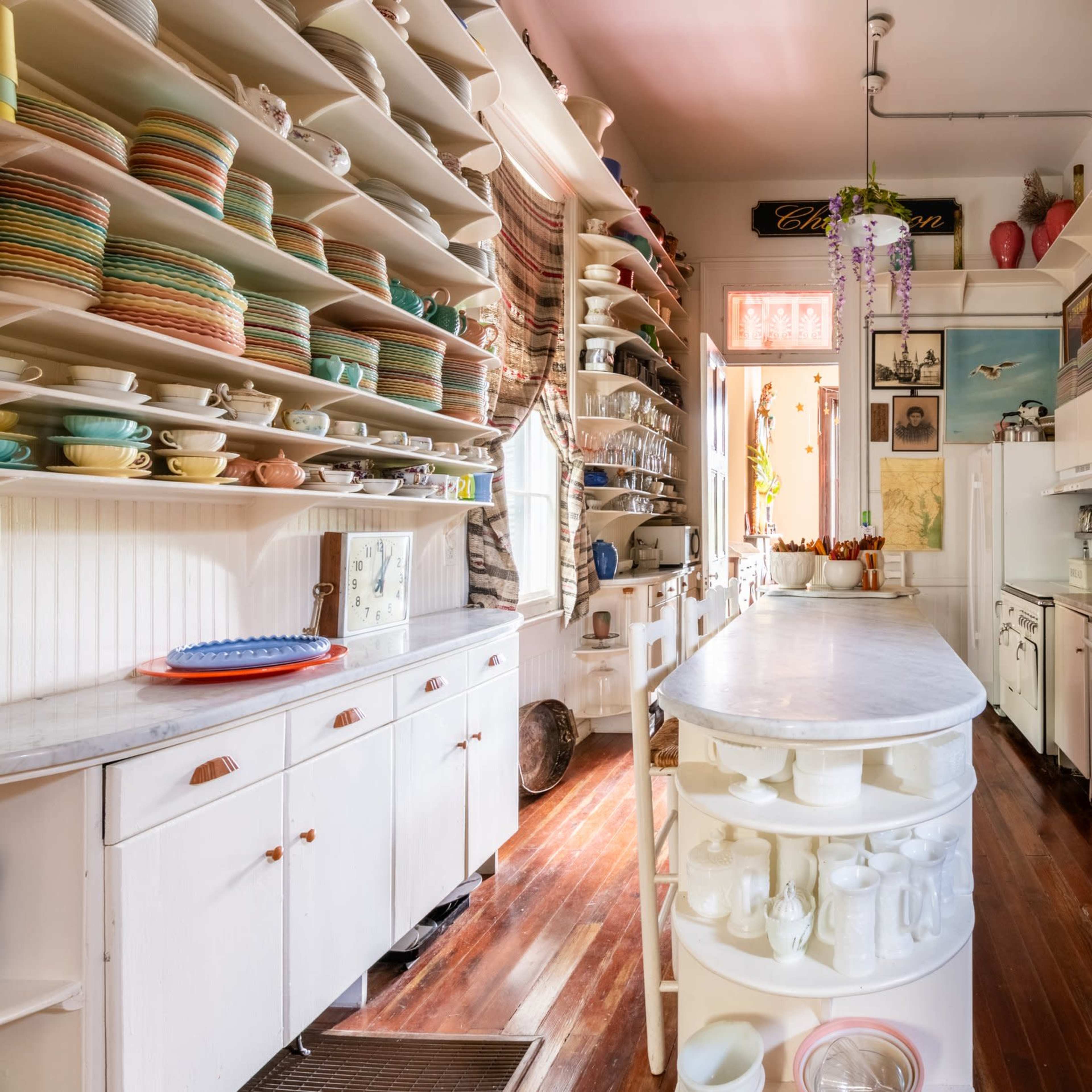A narrow, brightly lit kitchen lined with open shelves filled with colorful dishware and a central island with a white countertop.