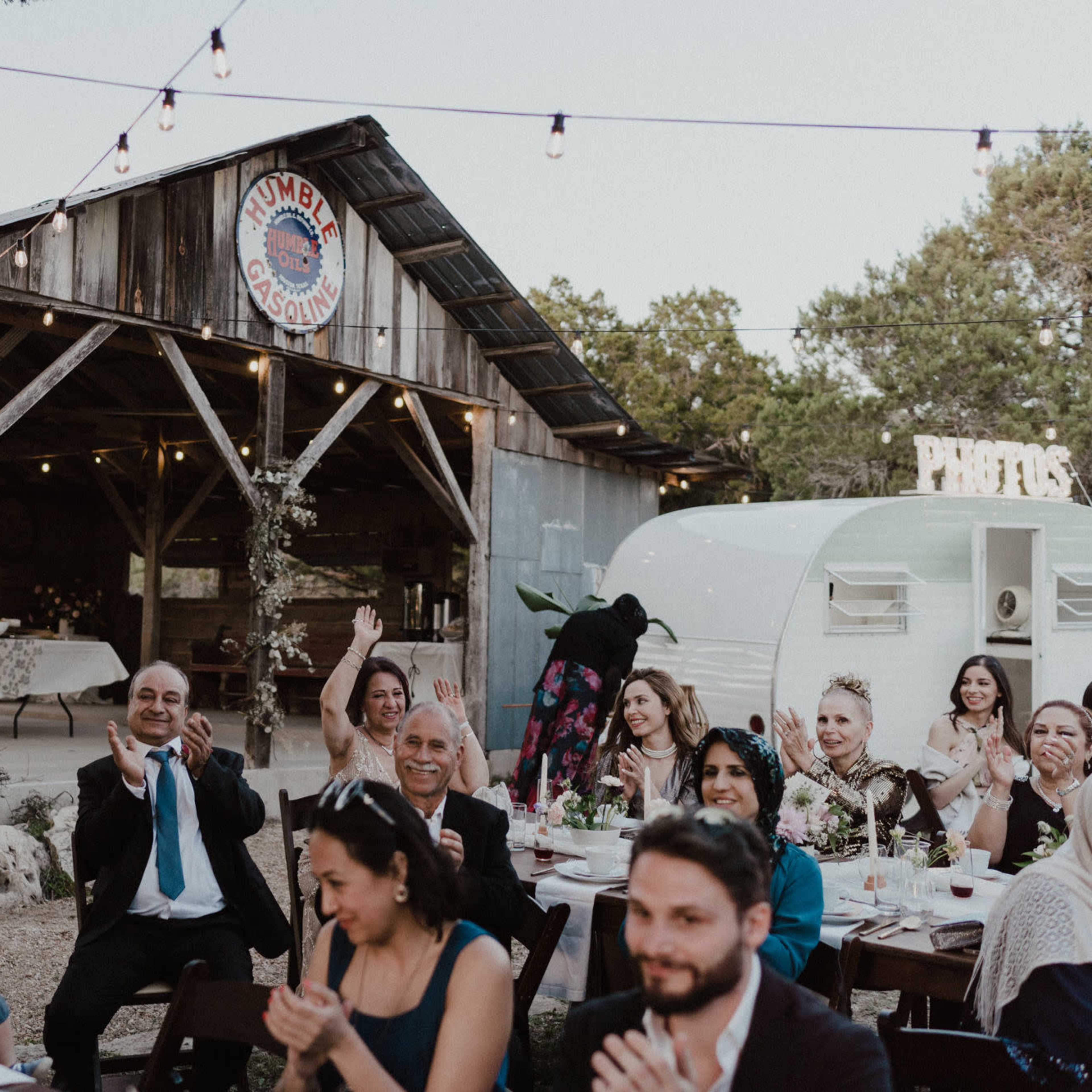 A group of guests applauds at a rustic outdoor celebration in front of a wooden barn adorned with string lights.