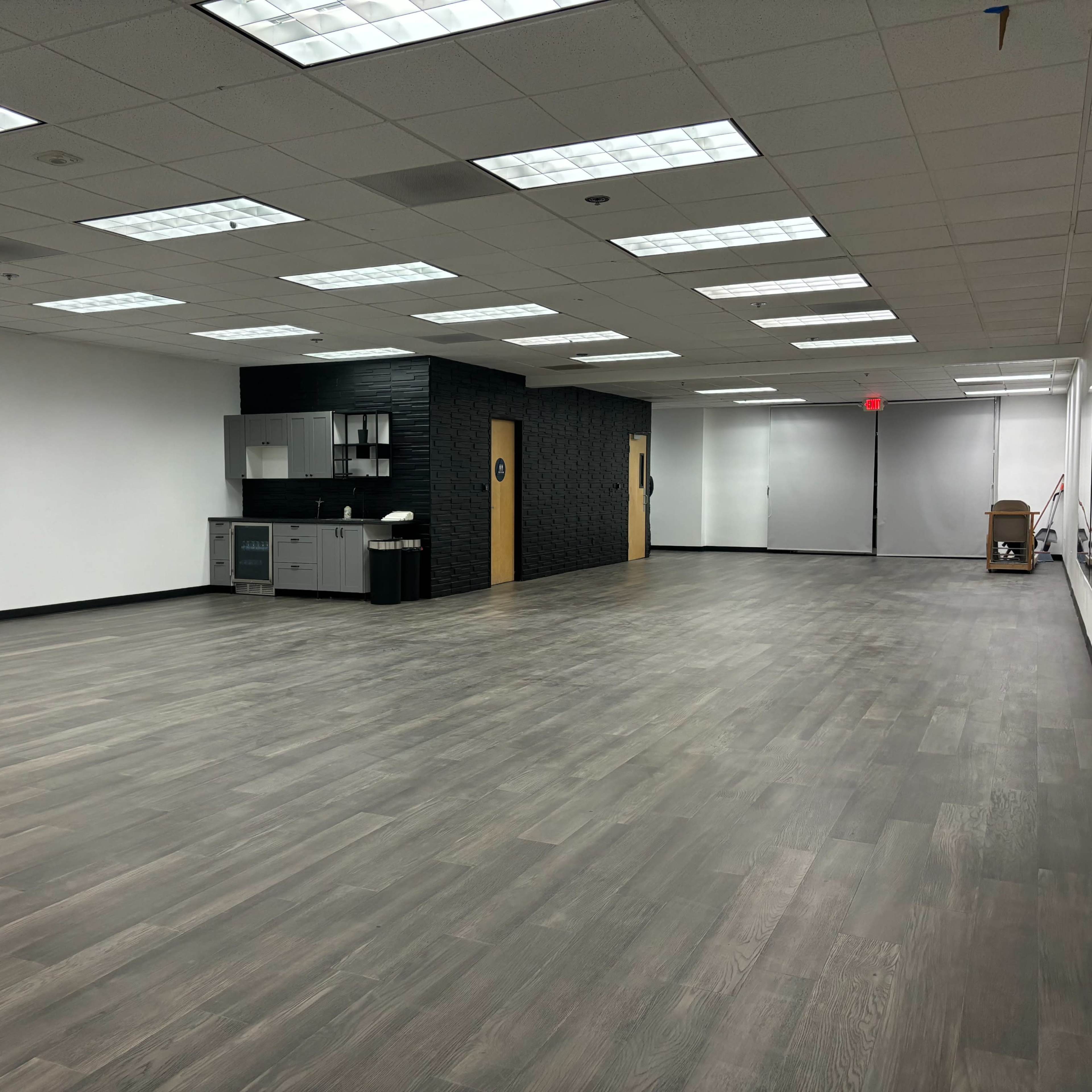 A spacious, empty room with wooden flooring, a kitchenette on one wall, and mirrors along another wall.