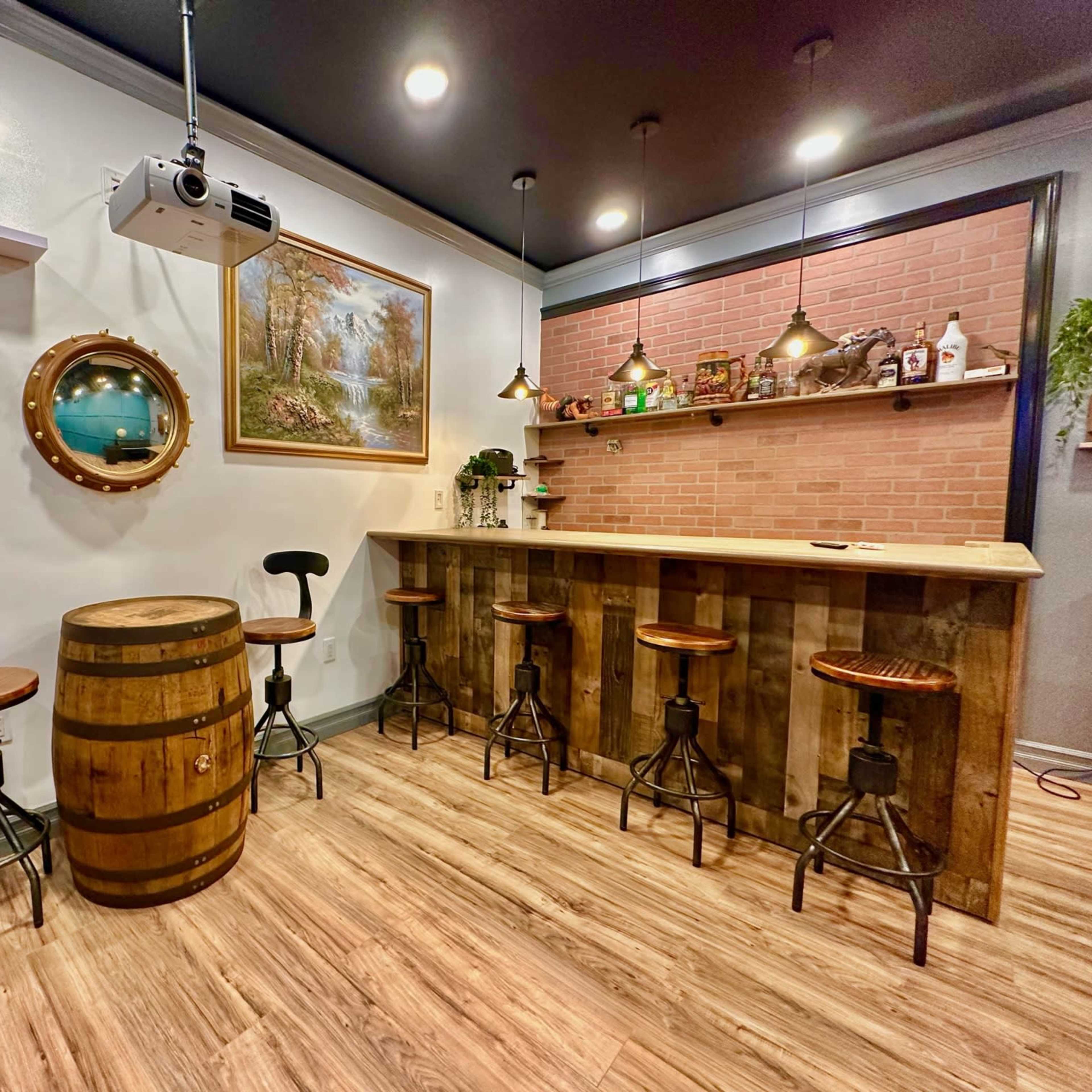 A cozy bar area with wooden stools surrounding a barrel table, a brick wall with shelves, and a projector mounted on the ceiling.