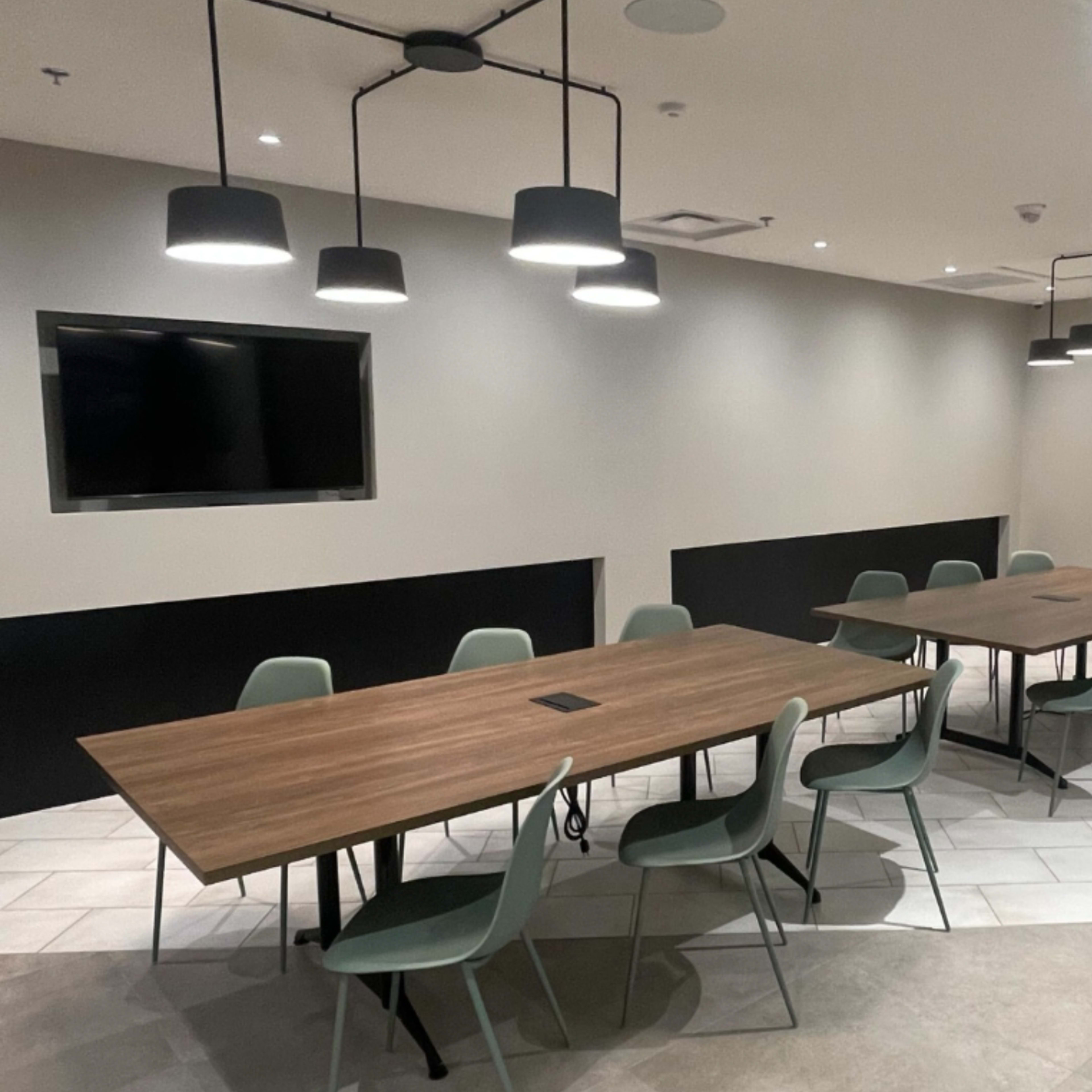 A modern conference room features two long wooden tables surrounded by green chairs, with pendant lights and a wall-mounted television.