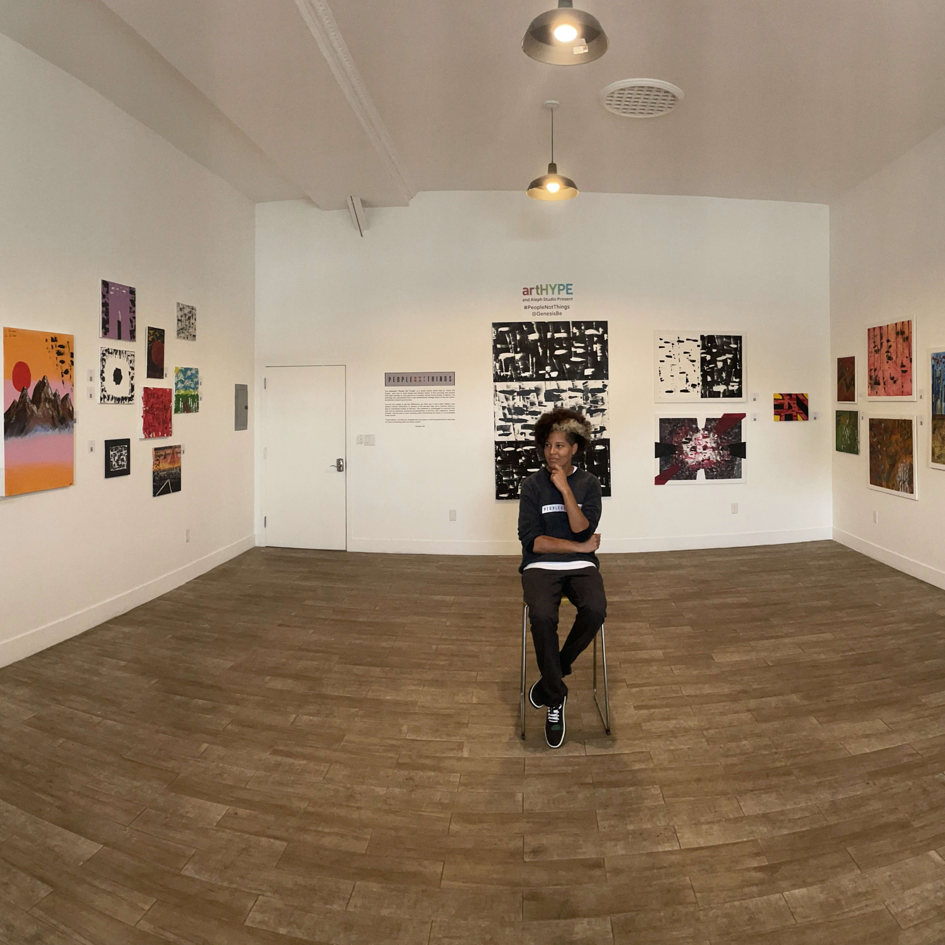 A person sits on a chair in the center of an art gallery surrounded by various artworks on the walls.