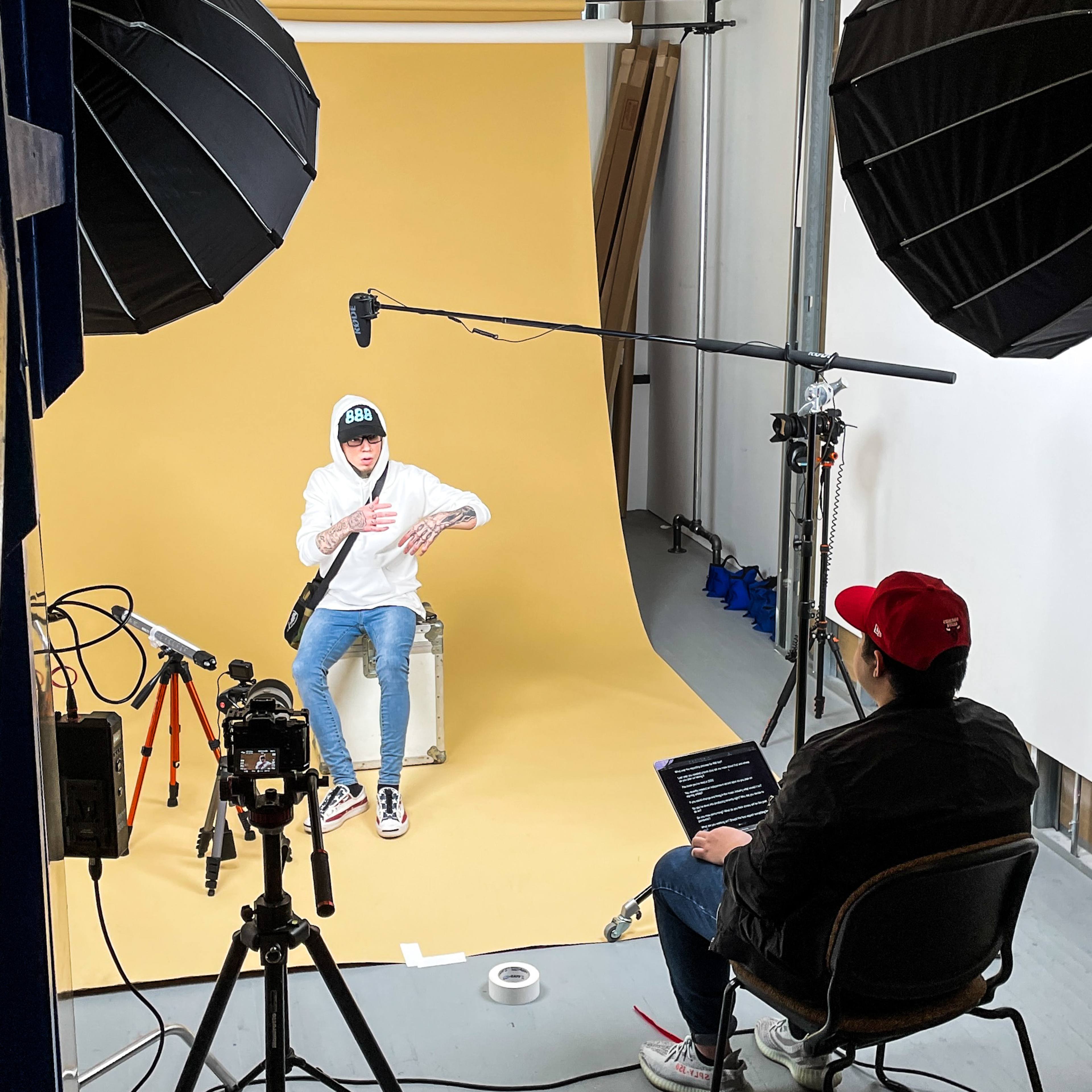 A young person wearing a hoodie is seated on a stool in front of a yellow backdrop, while another person in a red cap operates a laptop and records from a chair nearby.