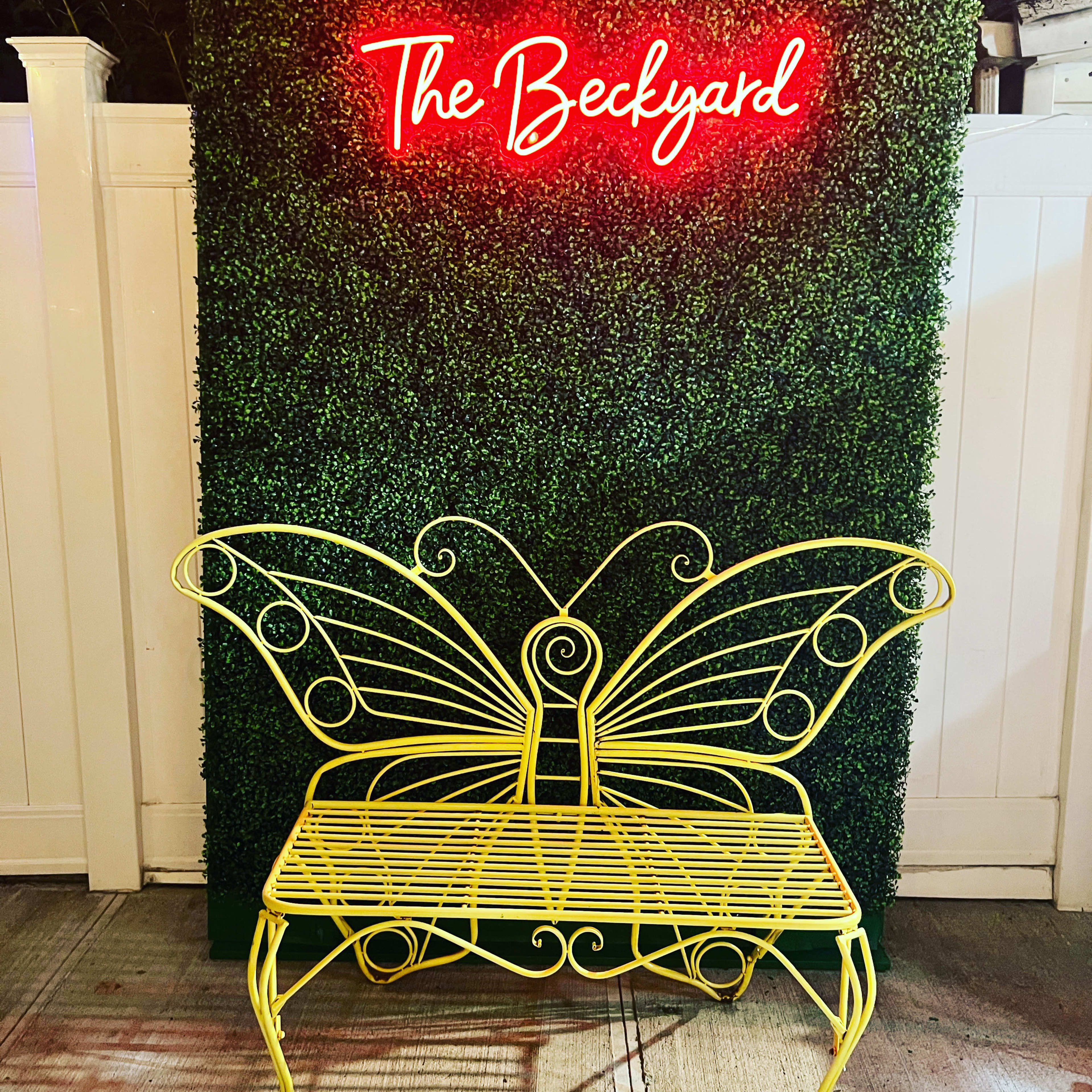 A yellow butterfly-shaped bench is positioned in front of a green hedge with a glowing red sign that reads "The Backyard."