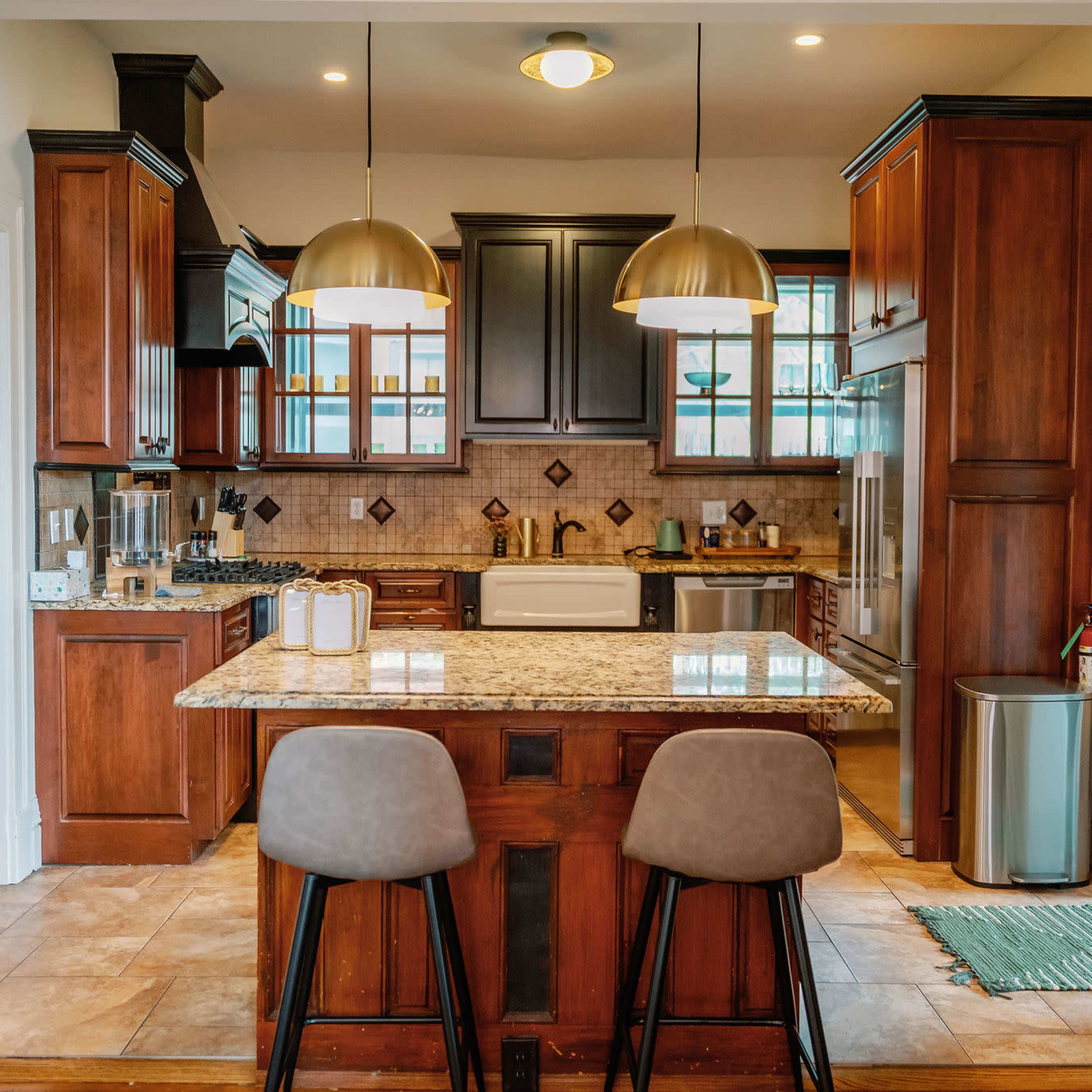 A modern kitchen features wooden cabinets, a large island with two stools, and pendant lighting above.