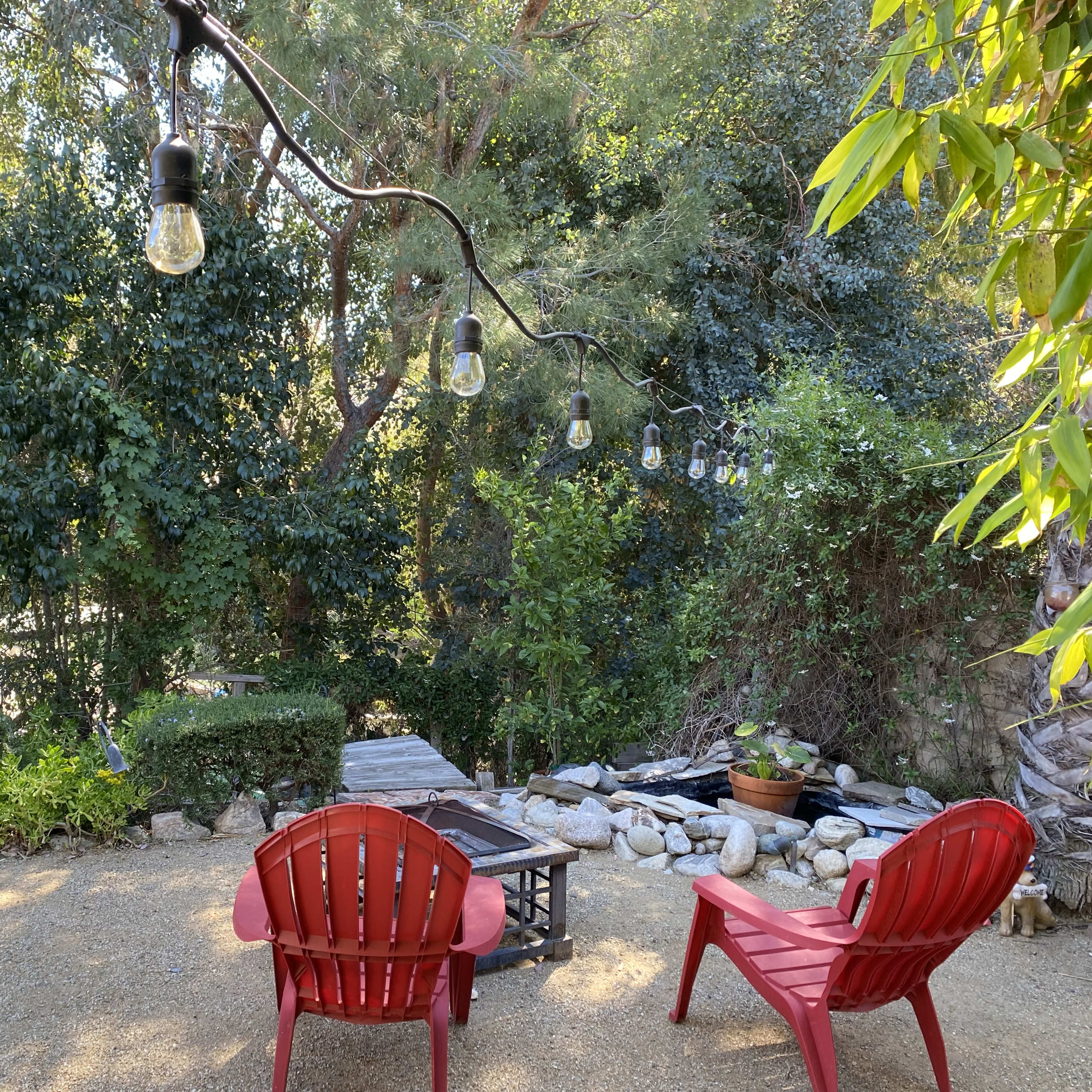 A backyard seating area features two red chairs, a small table, and string lights above, surrounded by greenery.