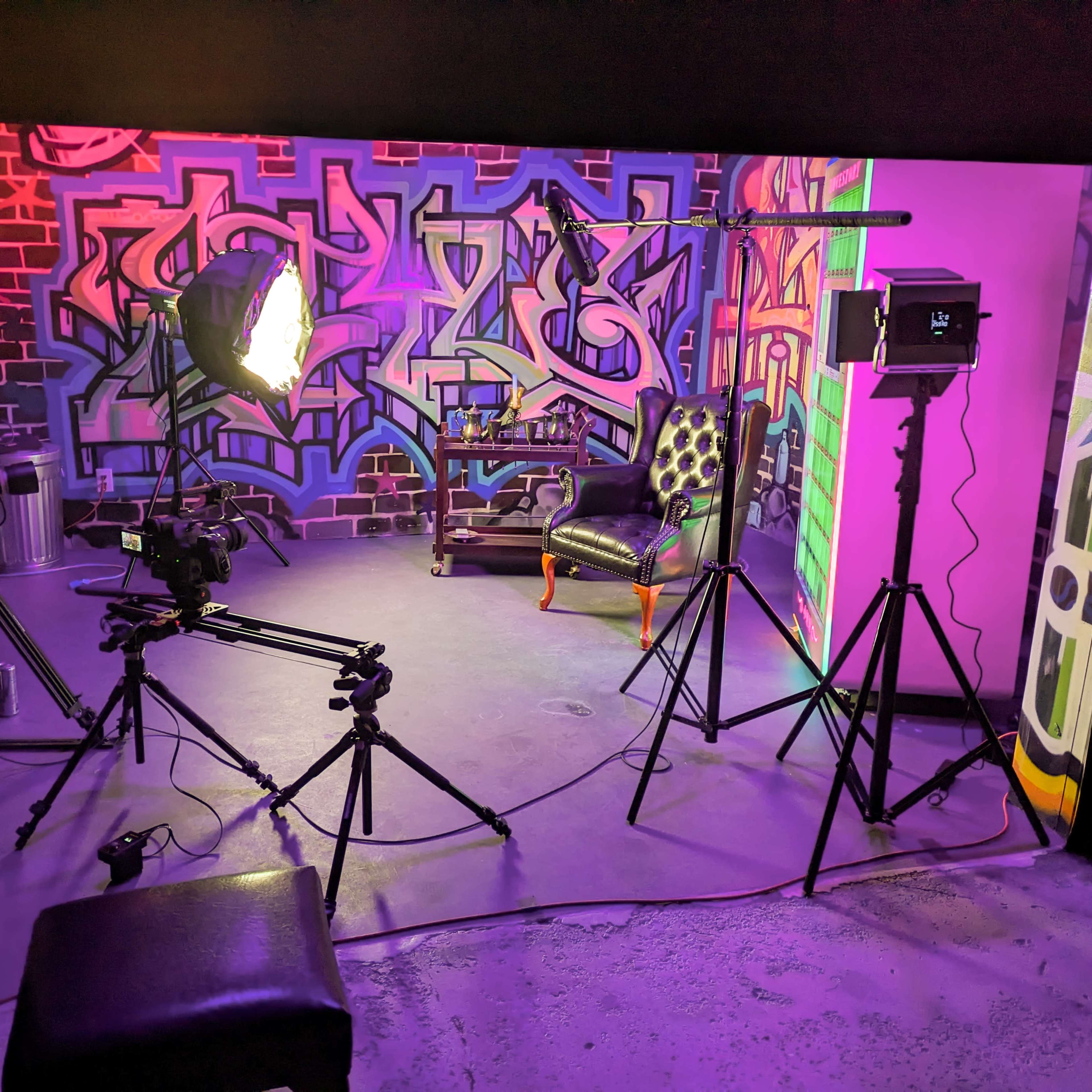 A photography studio setup with various lights and cameras positioned around a black chair in front of a colorful graffiti backdrop.