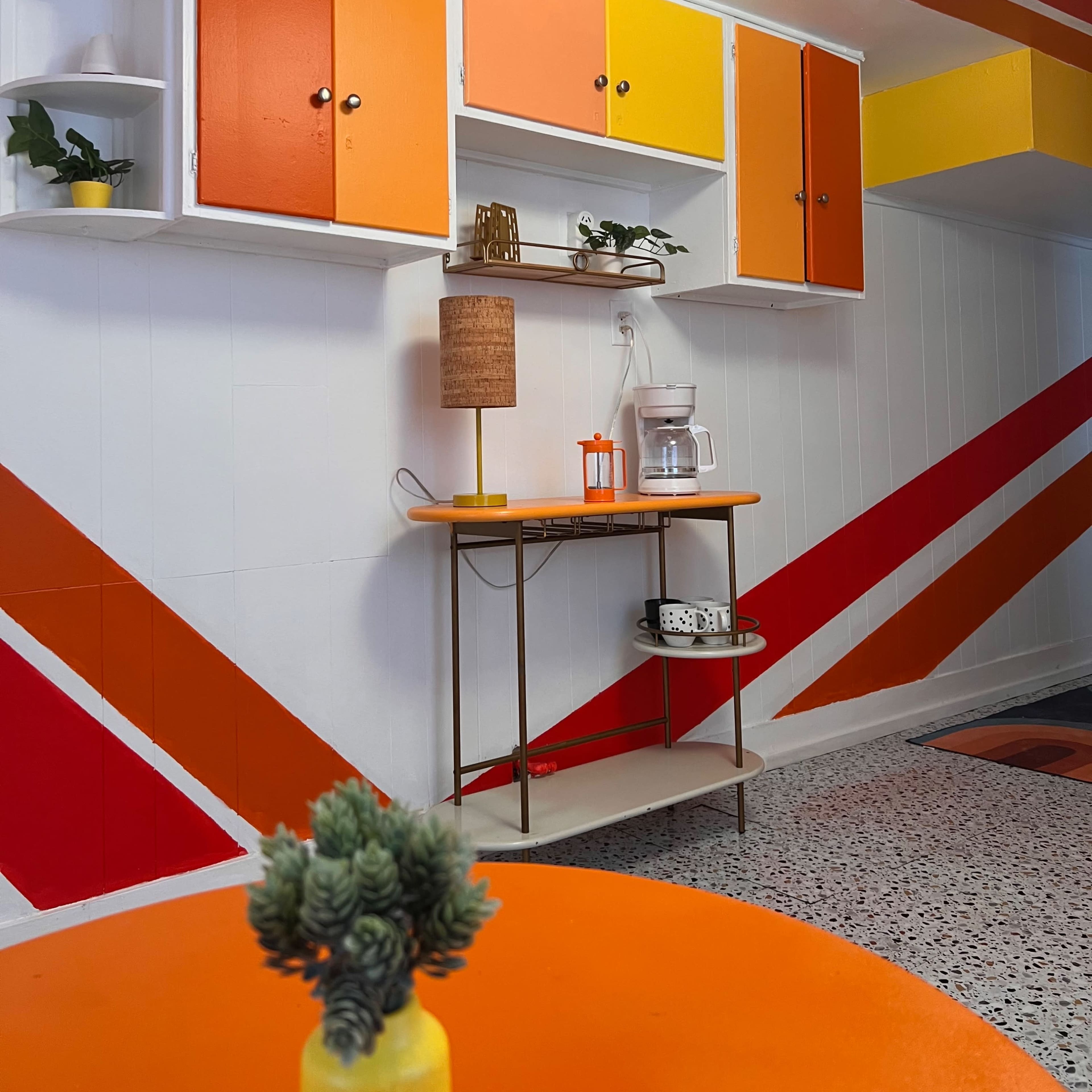A modern kitchen with orange and yellow cabinets and geometric wall patterns, featuring a small coffee station on a table.