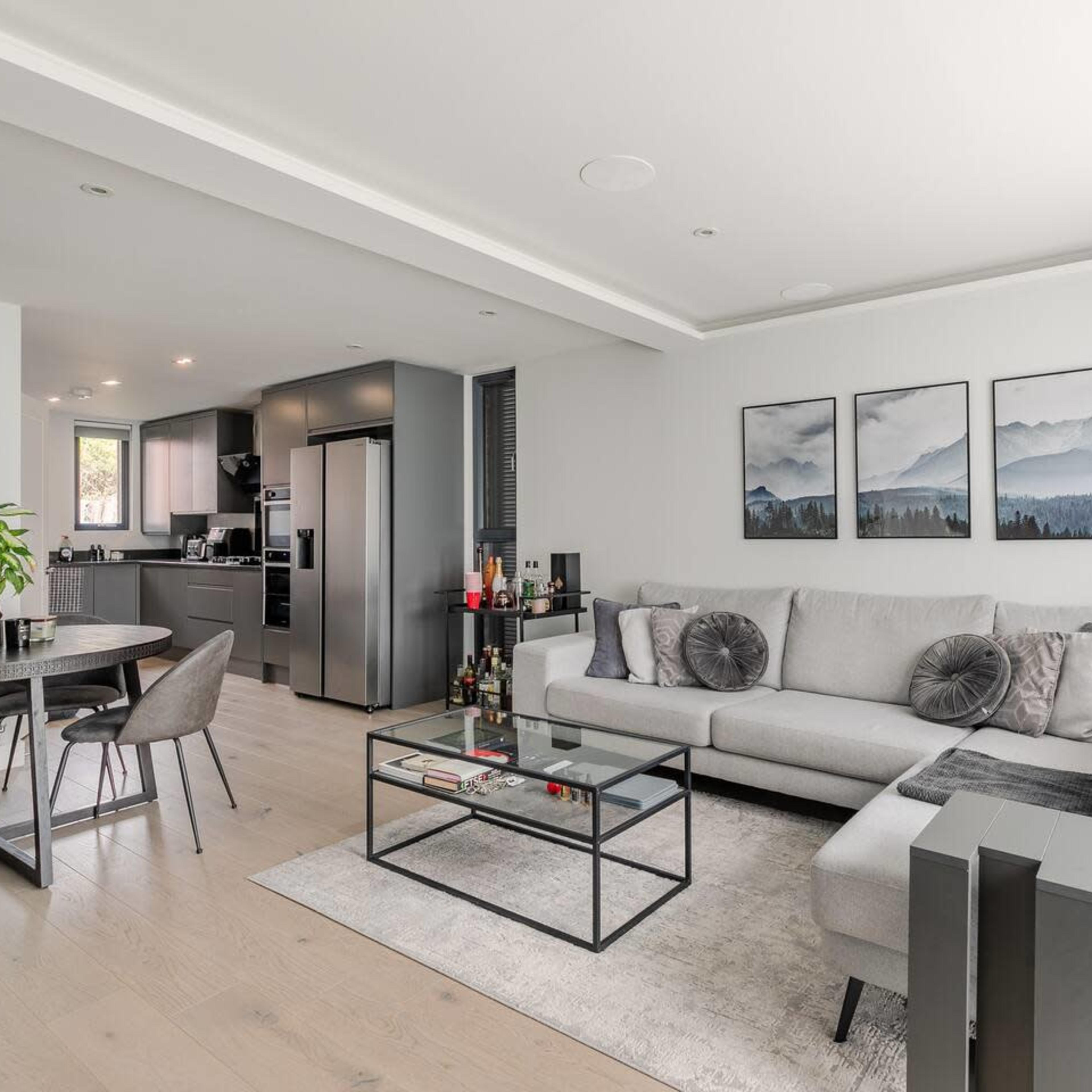 A modern living room featuring a gray sectional sofa, a glass coffee table, and a dining area with a round table and chairs, all set against a backdrop of a sleek kitchen.