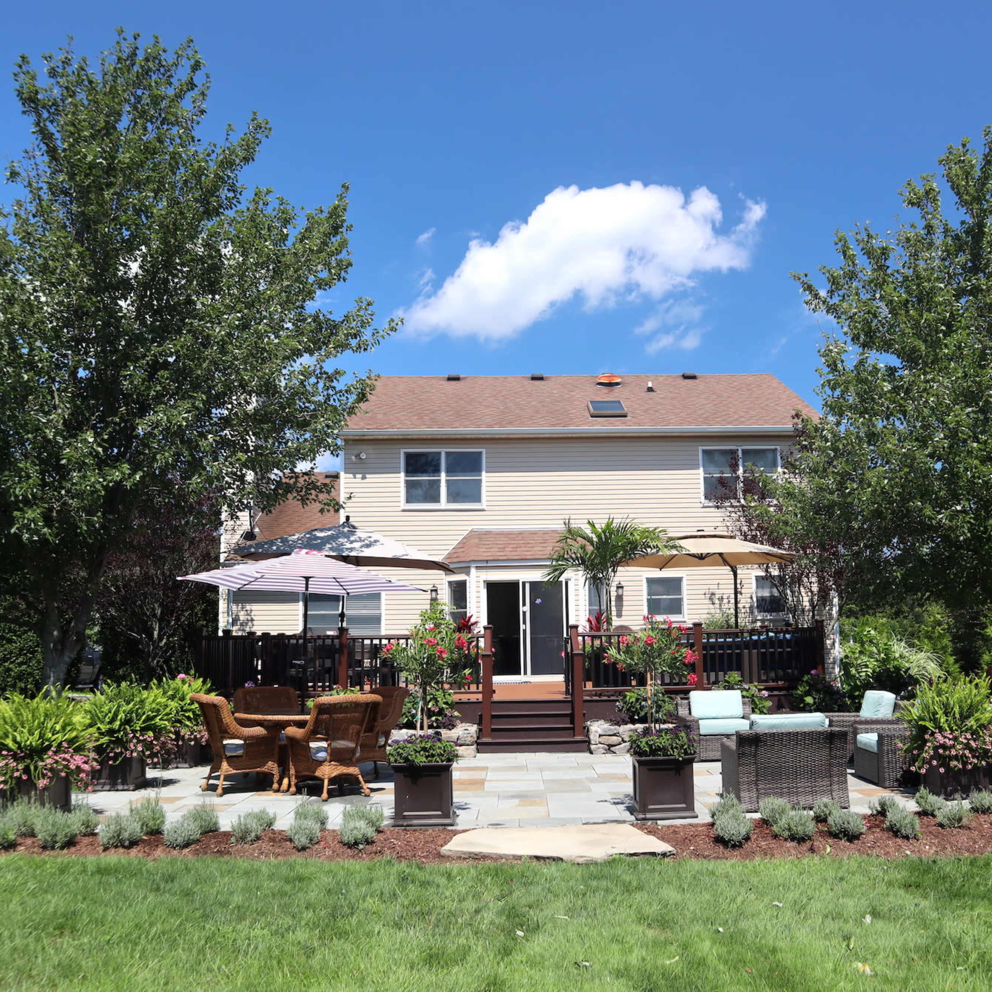 A two-story house with a backyard patio area featuring seating, landscaping, and trees under a blue sky.