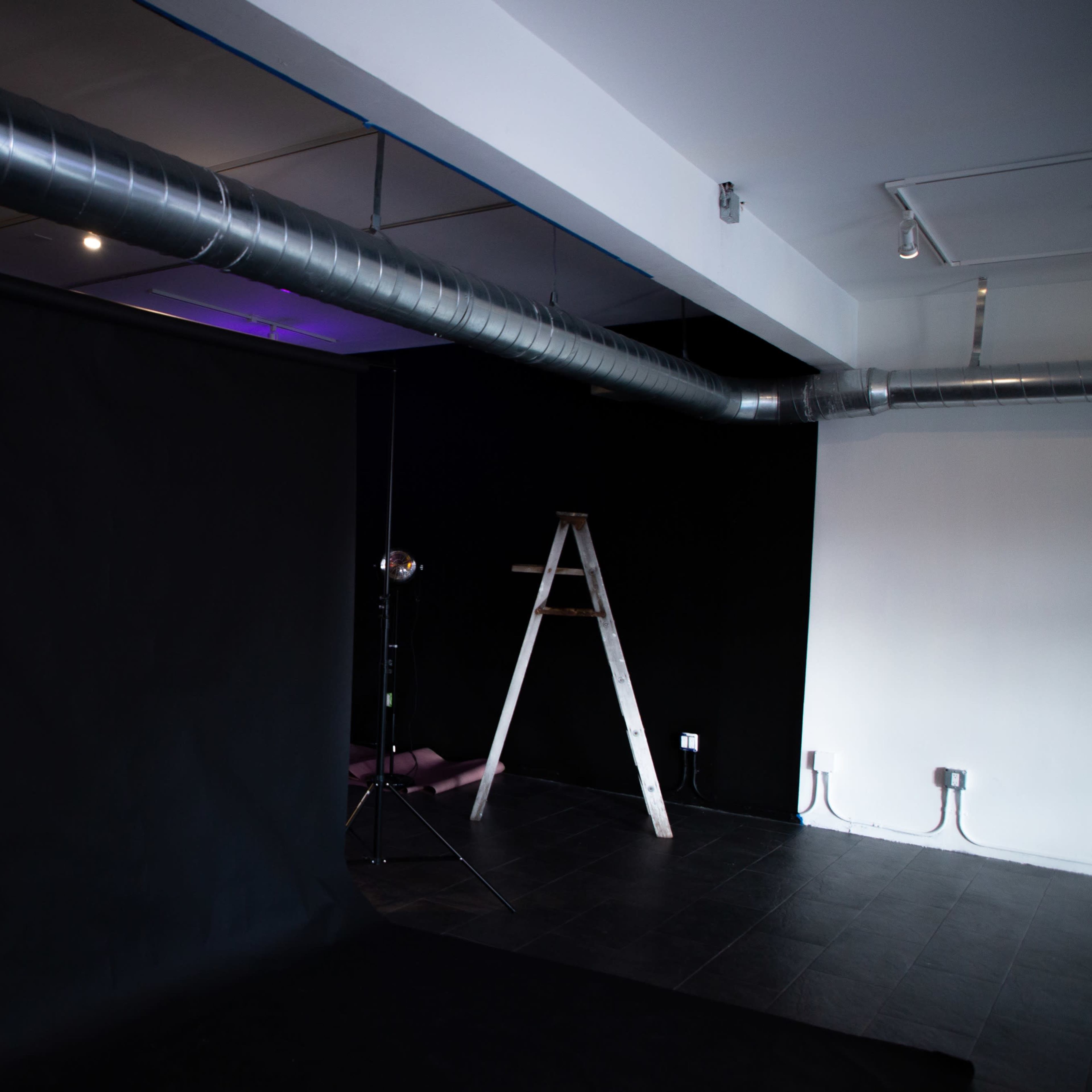A photo studio with a black backdrop, a ladder, and exposed metal ductwork against white walls.