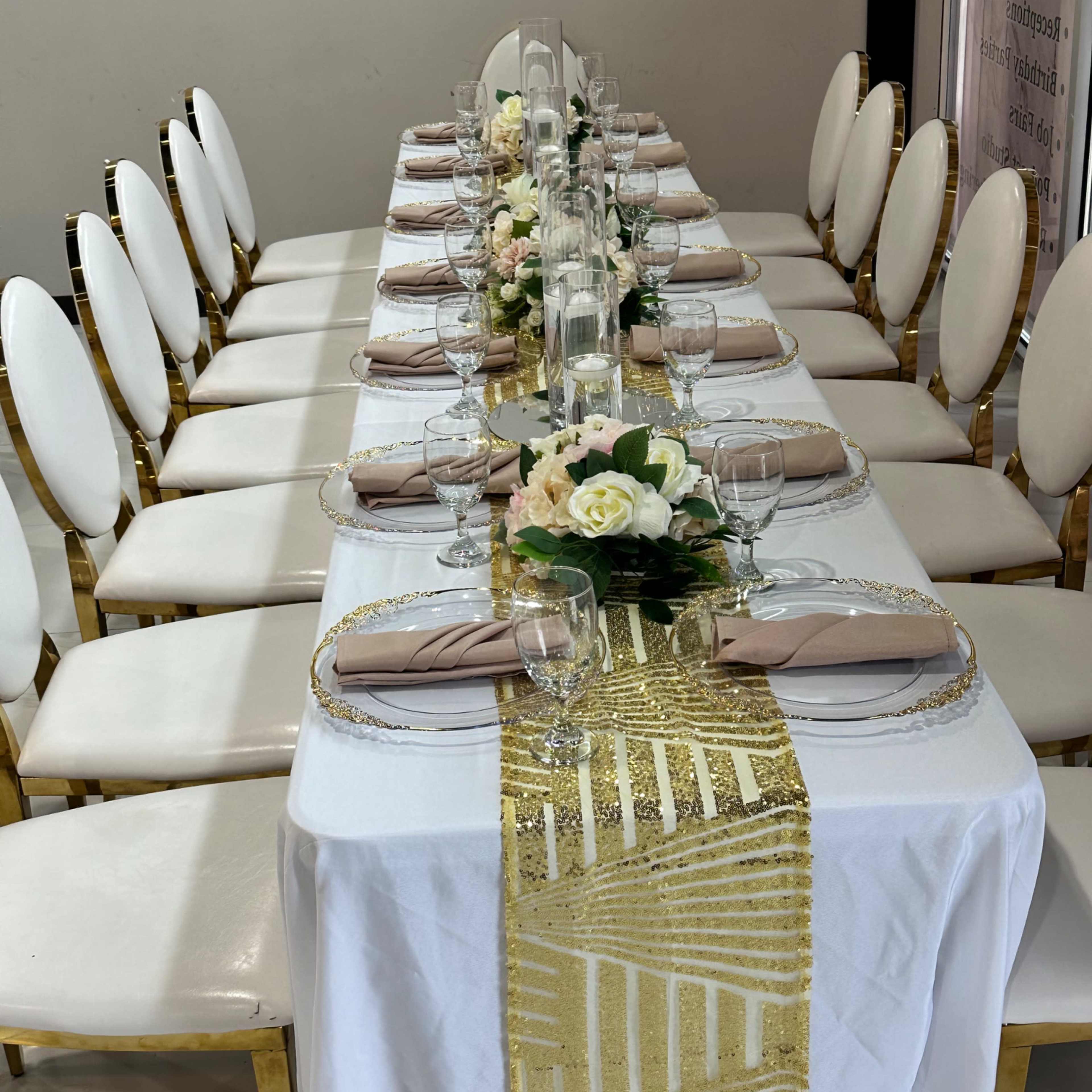 A long dining table is set with white linens, gold accents, and floral centerpieces, lined with elegant chairs.