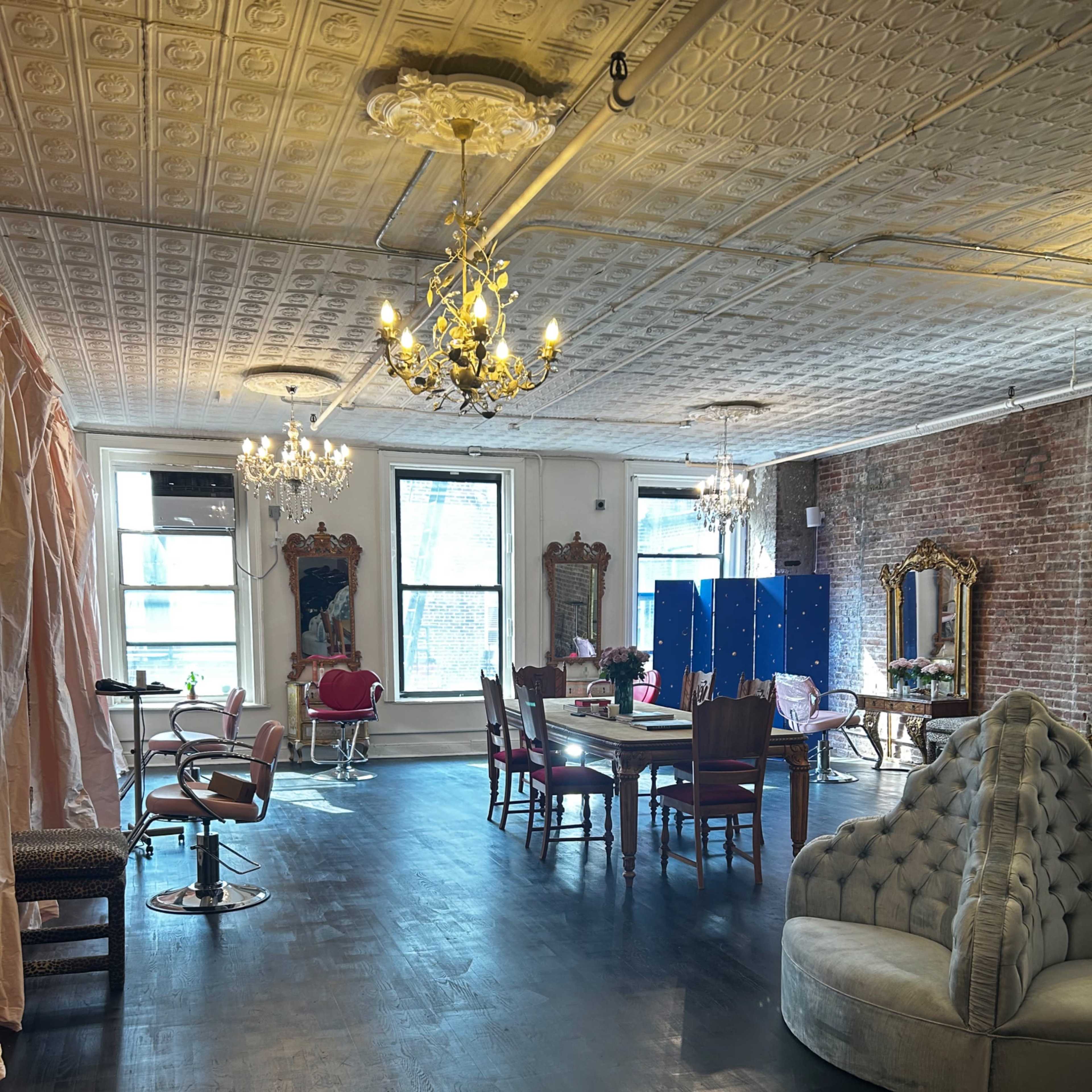 A spacious room with vintage-style furniture, chandeliers, and large windows, featuring a mixture of seating areas and a dining table.
