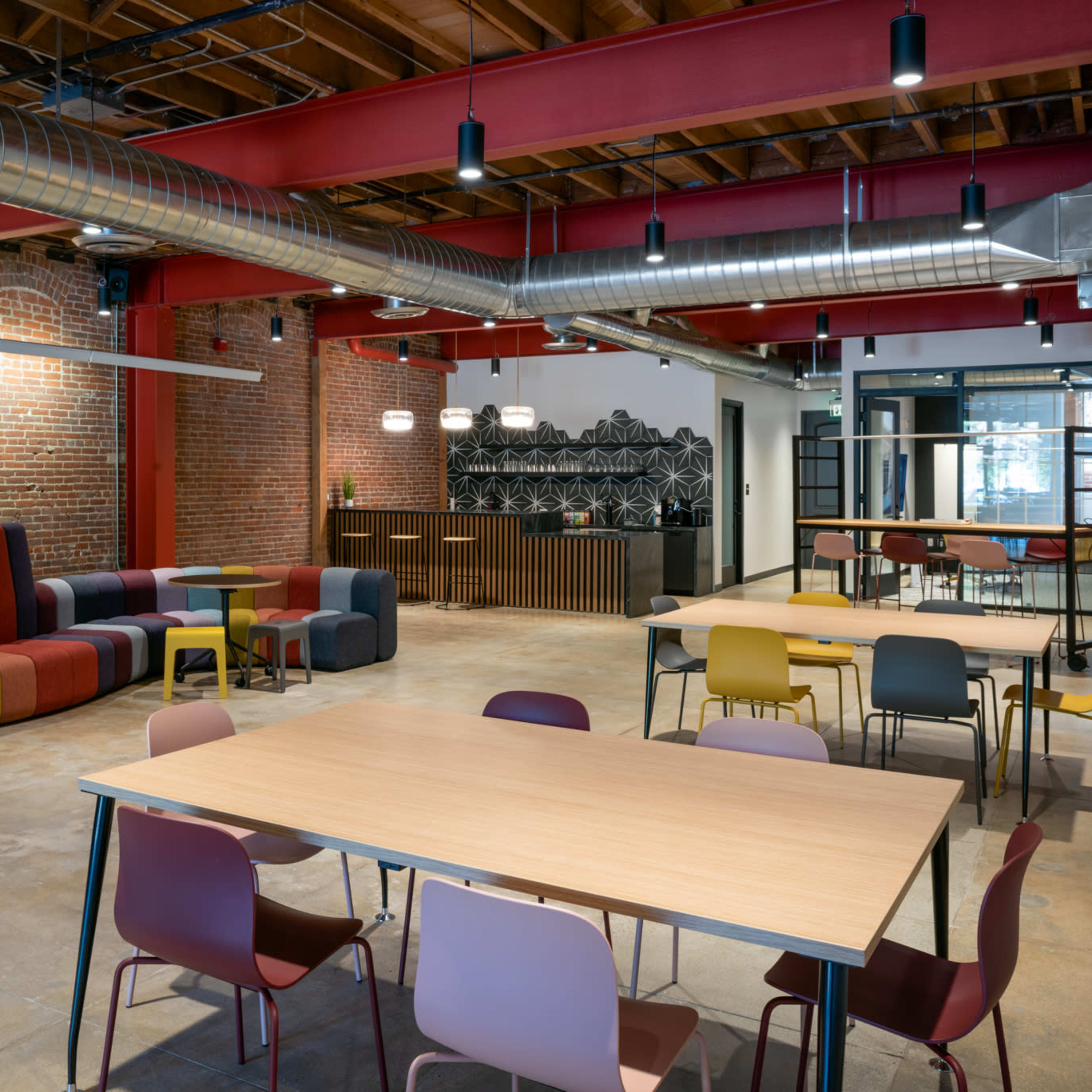 A modern workspace featuring colorful seating, wooden tables, and exposed brick walls with industrial-style lighting.