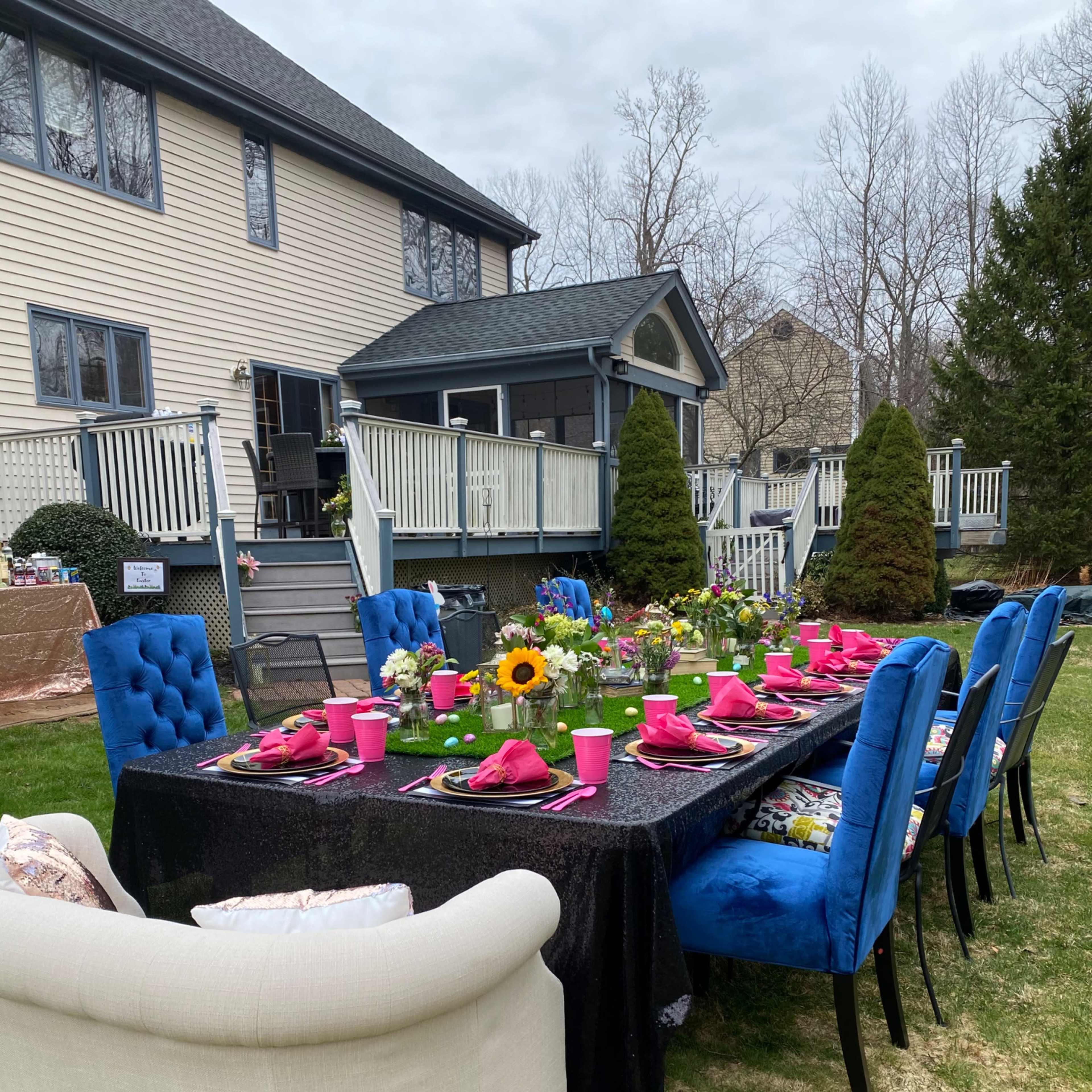 A large outdoor dining table is set for a meal, decorated with flowers and surrounded by chairs, with a house and yard in the background.
