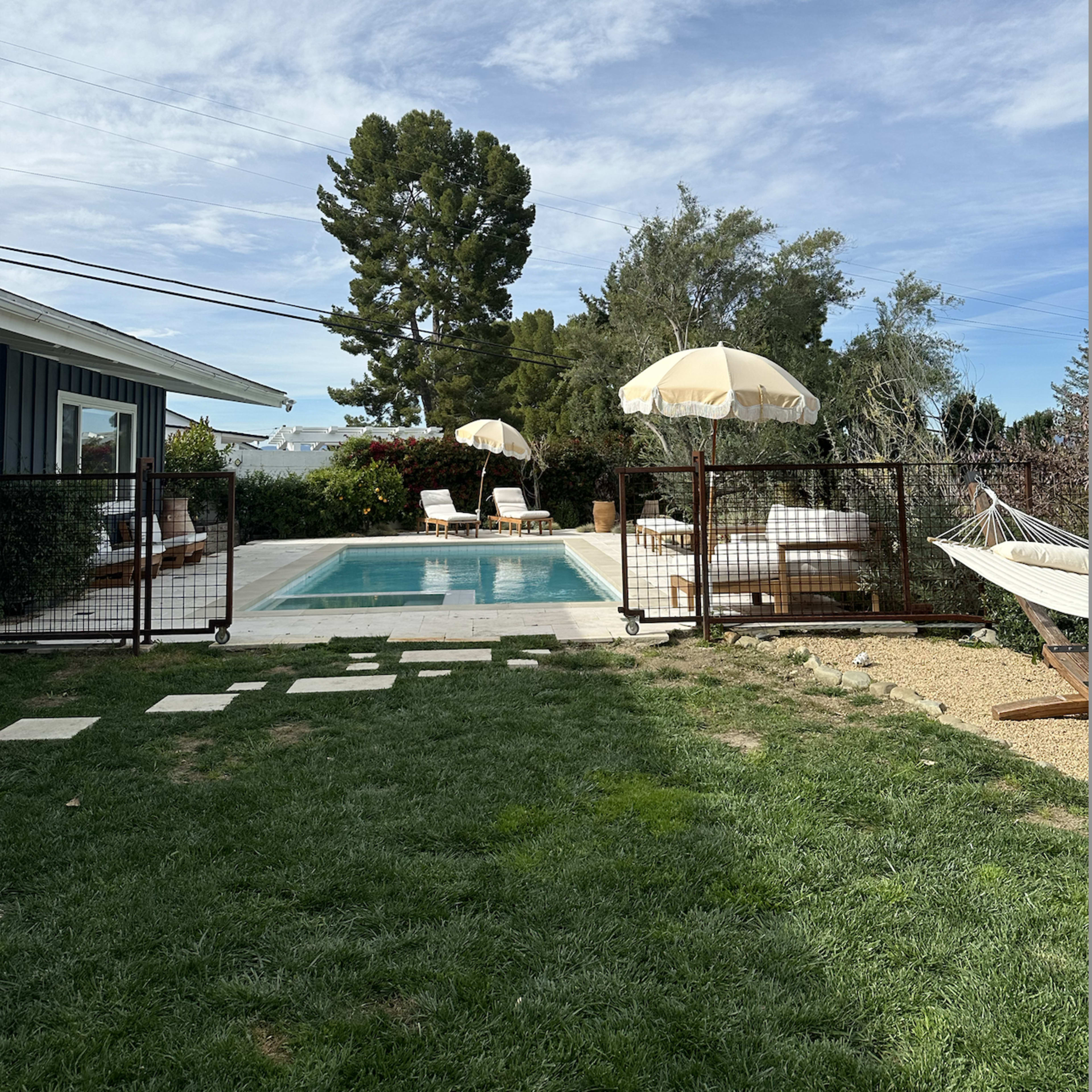 A rectangular pool is surrounded by lounge chairs and umbrellas in a backyard, with a hammock nearby and trees in the background.
