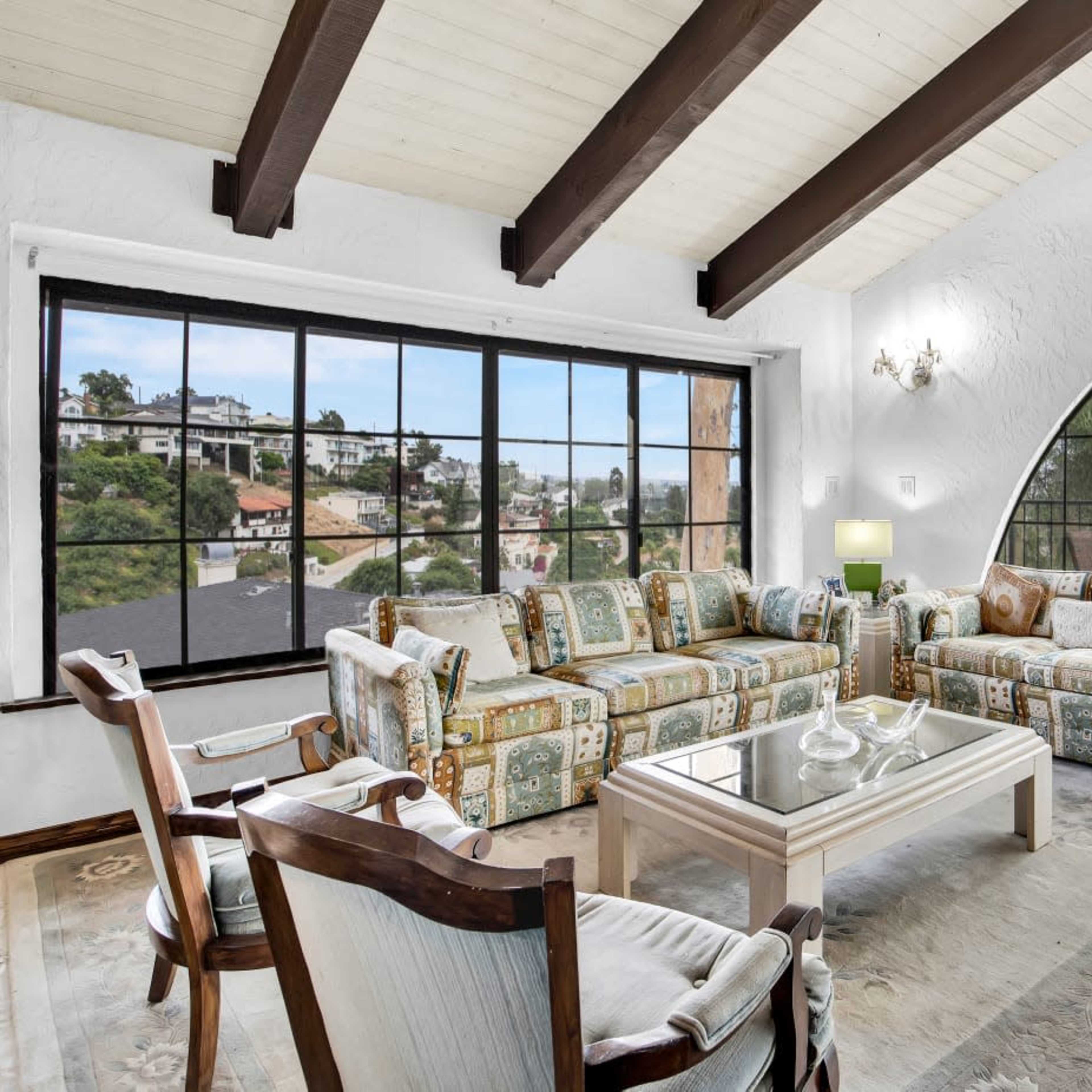 A spacious living room features arched windows with a view, wooden beams on the ceiling, and a mix of patterned furniture arranged around a glass coffee table.