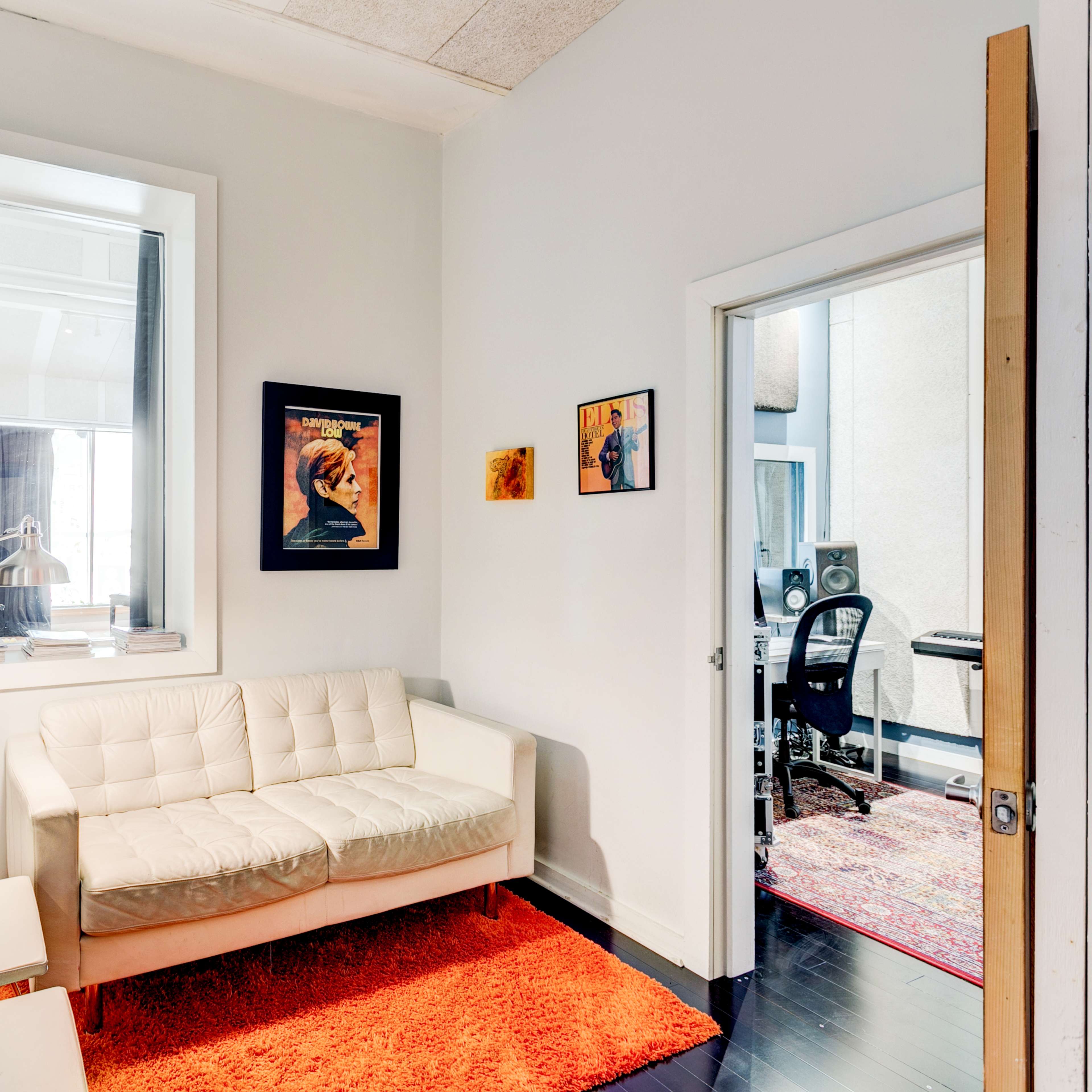 A small room with a white sofa, an orange rug, and framed artwork on the walls, leading to another room with studio equipment visible through an open door.
