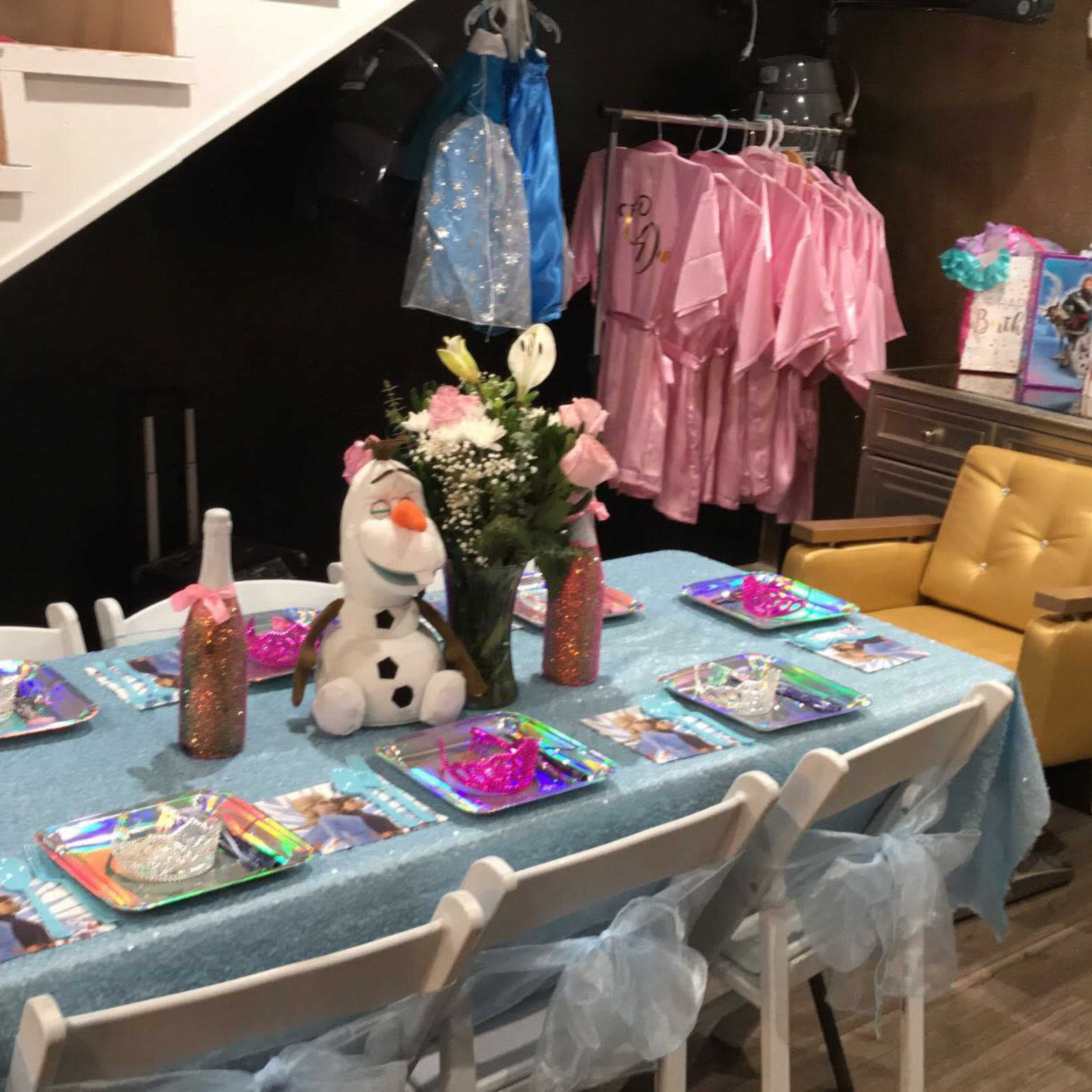 A dining area is set up with a blue tablecloth, decorated plates, and centerpieces, alongside pink dresses hanging on the wall and a staircase in the background.