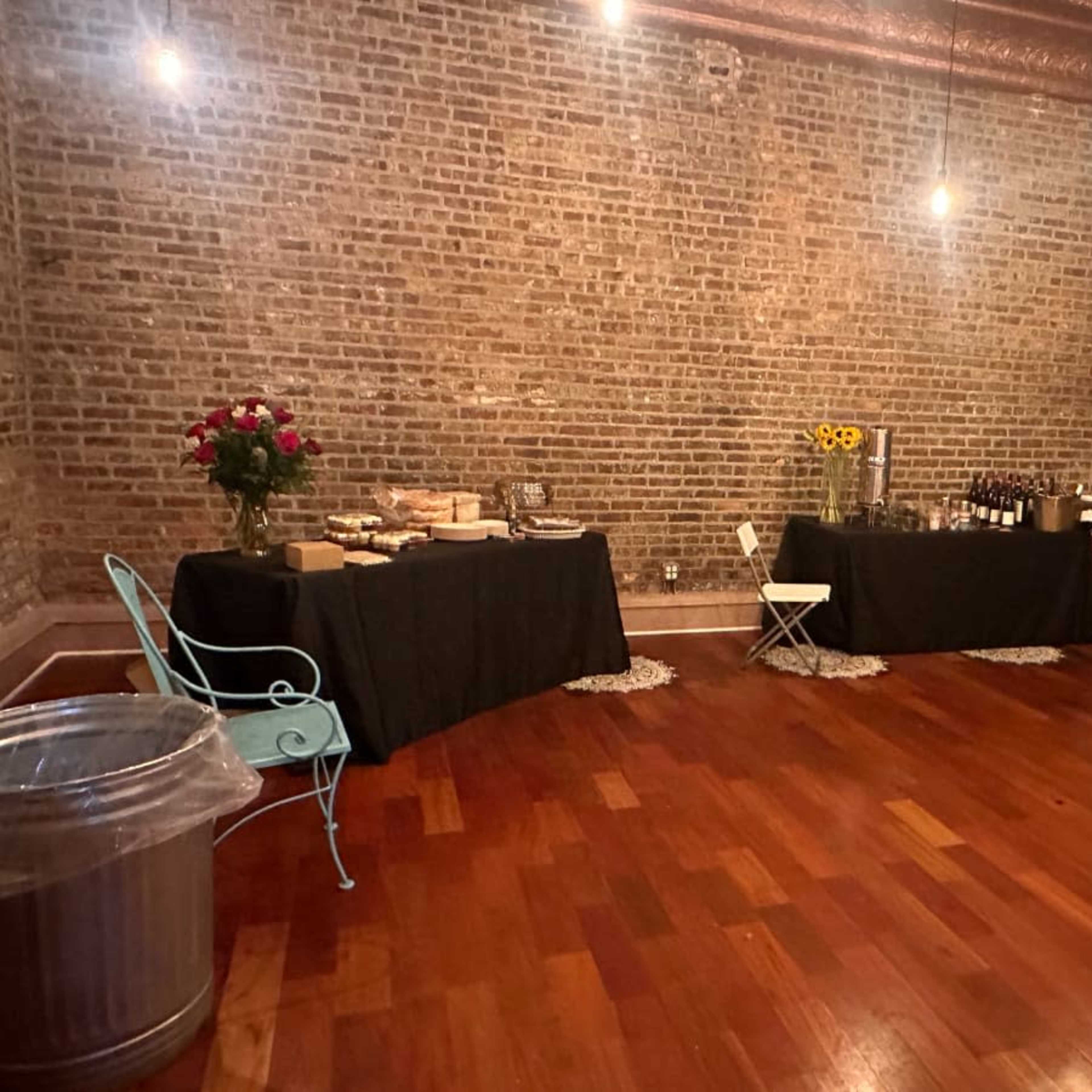 A decorated room with brick walls featuring two tables covered in black cloth, set with food and drinks, and a small blue chair nearby.