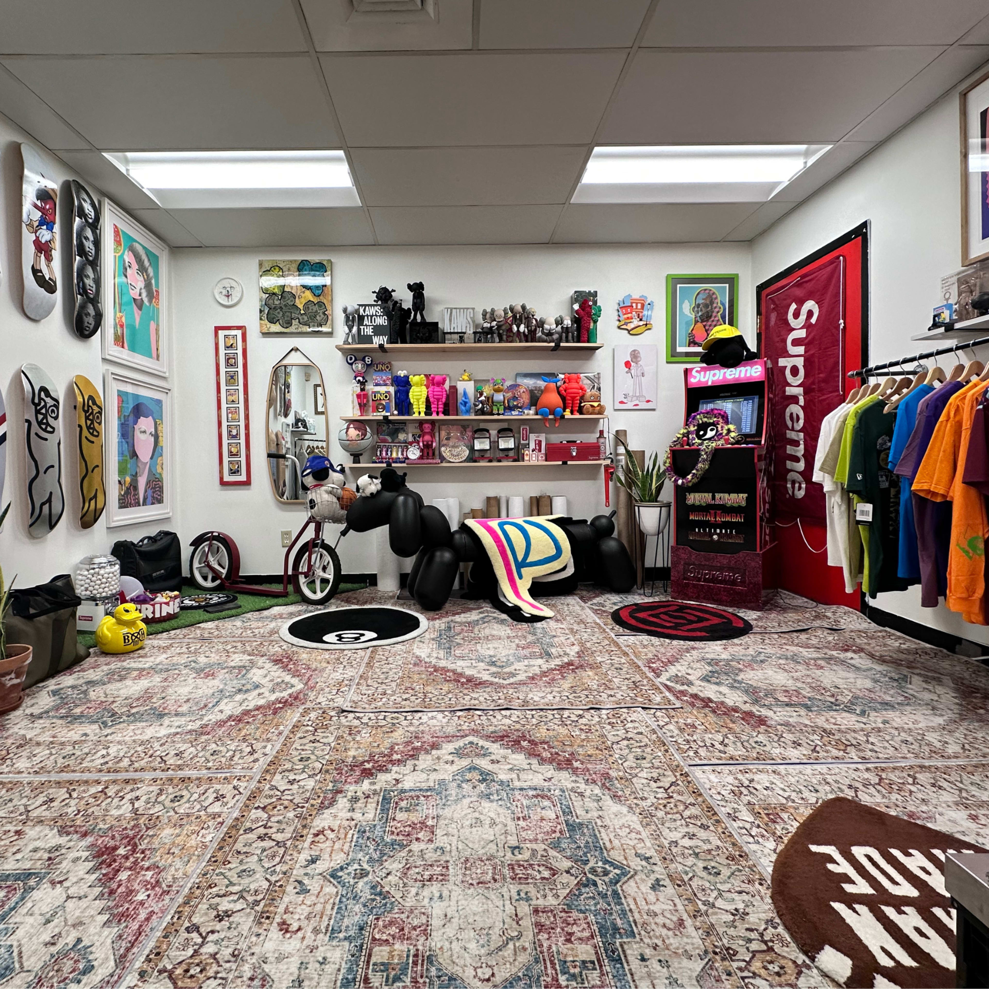 The room features a collection of colorful clothing, art pieces, and decorative items, with a patterned rug and several skateboards displayed on the walls.