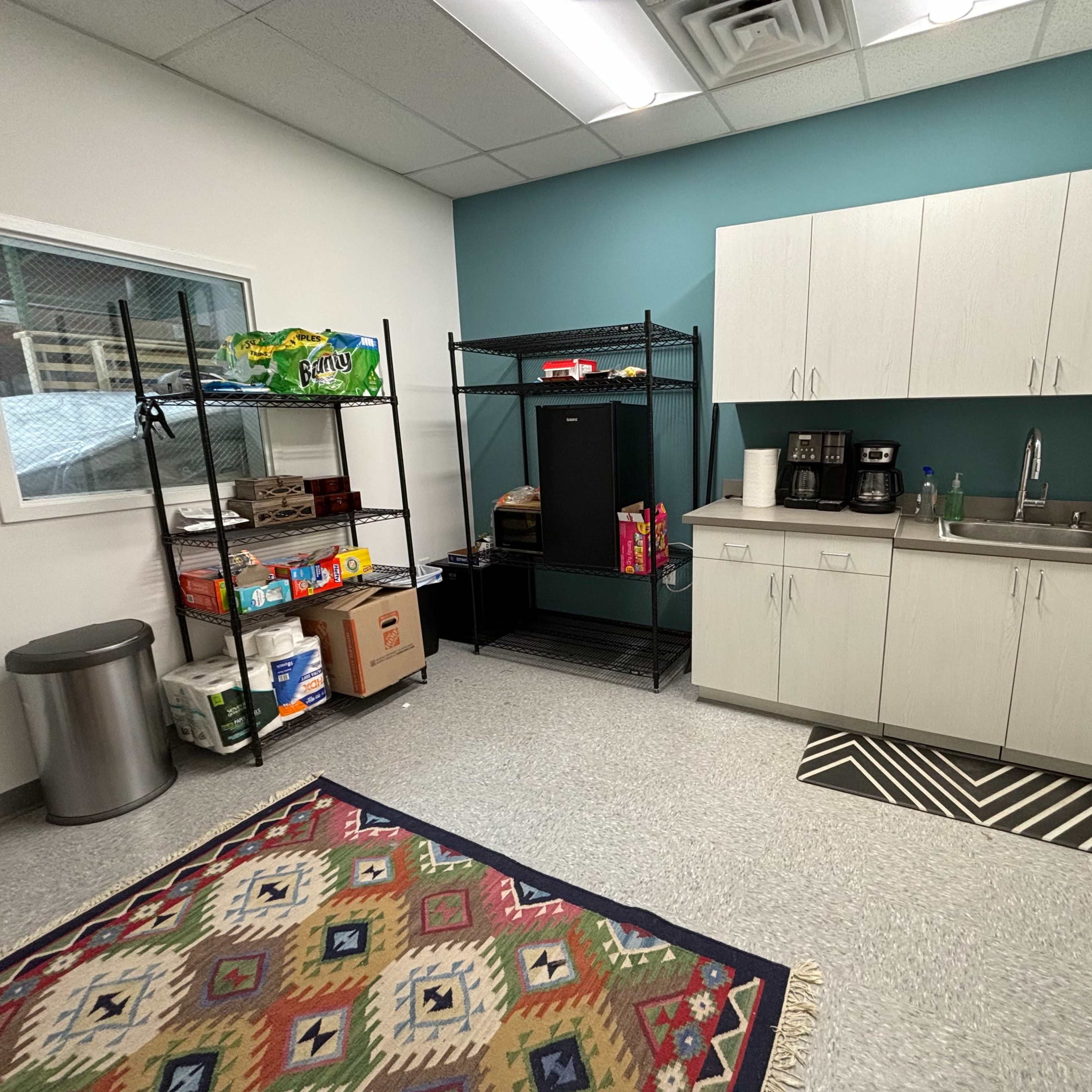 A small break room featuring a kitchenette with cabinets, a sink, and a coffee maker, along with a shelf stocked with snacks and beverages, and a patterned rug on the floor.