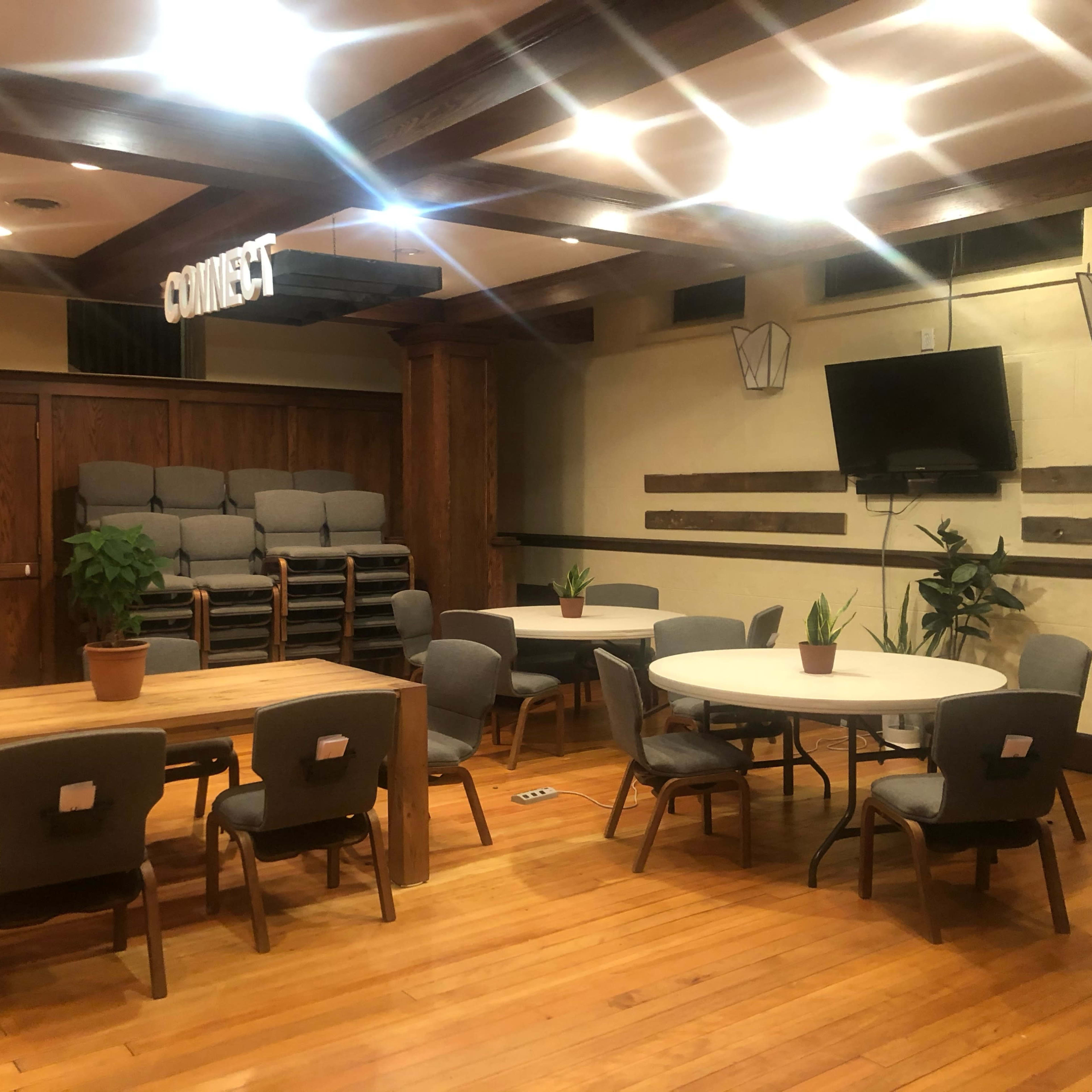 The room features several tables and chairs arranged for group seating, with a stacked set of gray chairs in the background and plants positioned on tables.