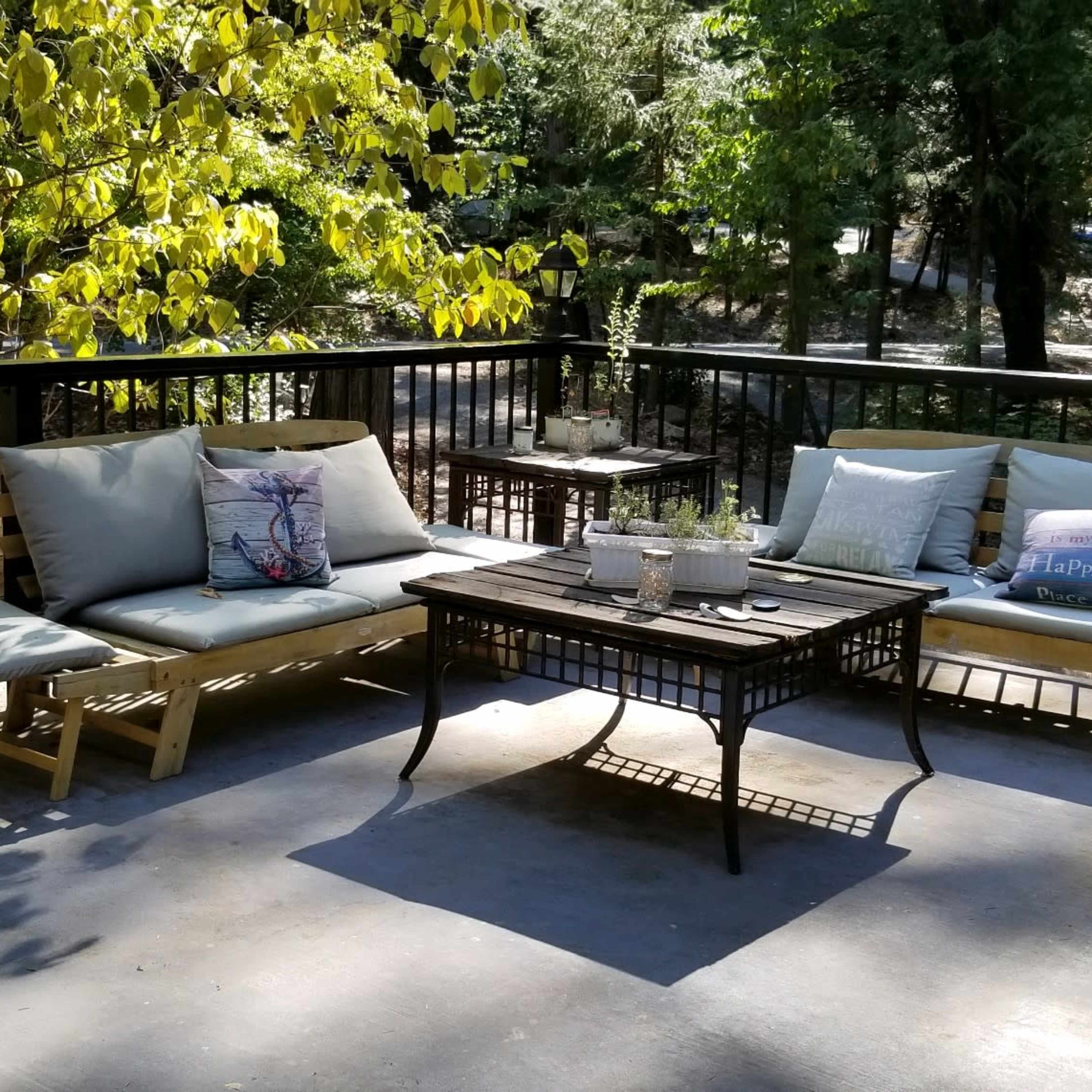 A covered outdoor patio features a light wooden sofa and dark metal tables surrounded by greenery.