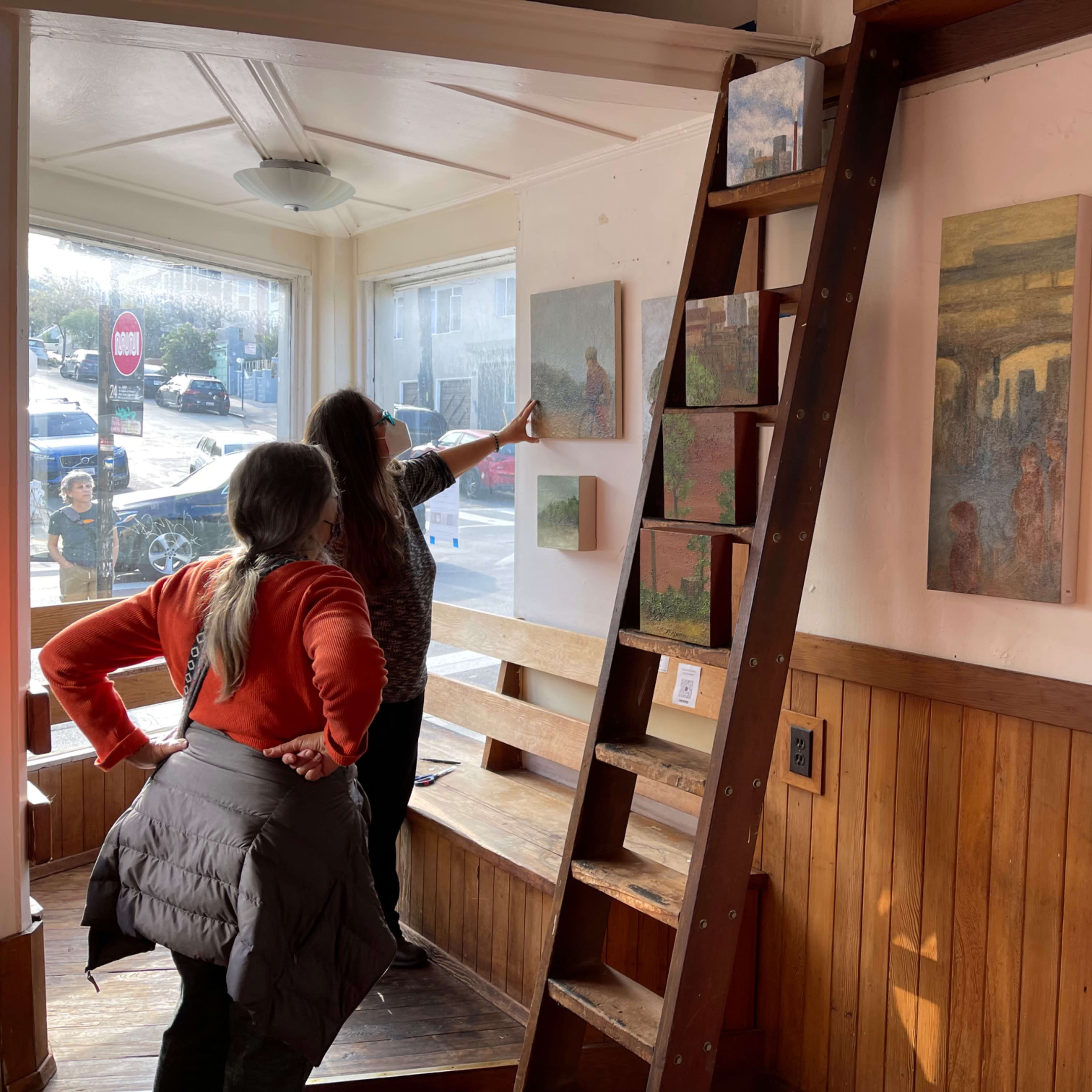Two women examine artwork on the walls of a gallery, while a wooden ladder leans against one wall and a street view is visible through the window.