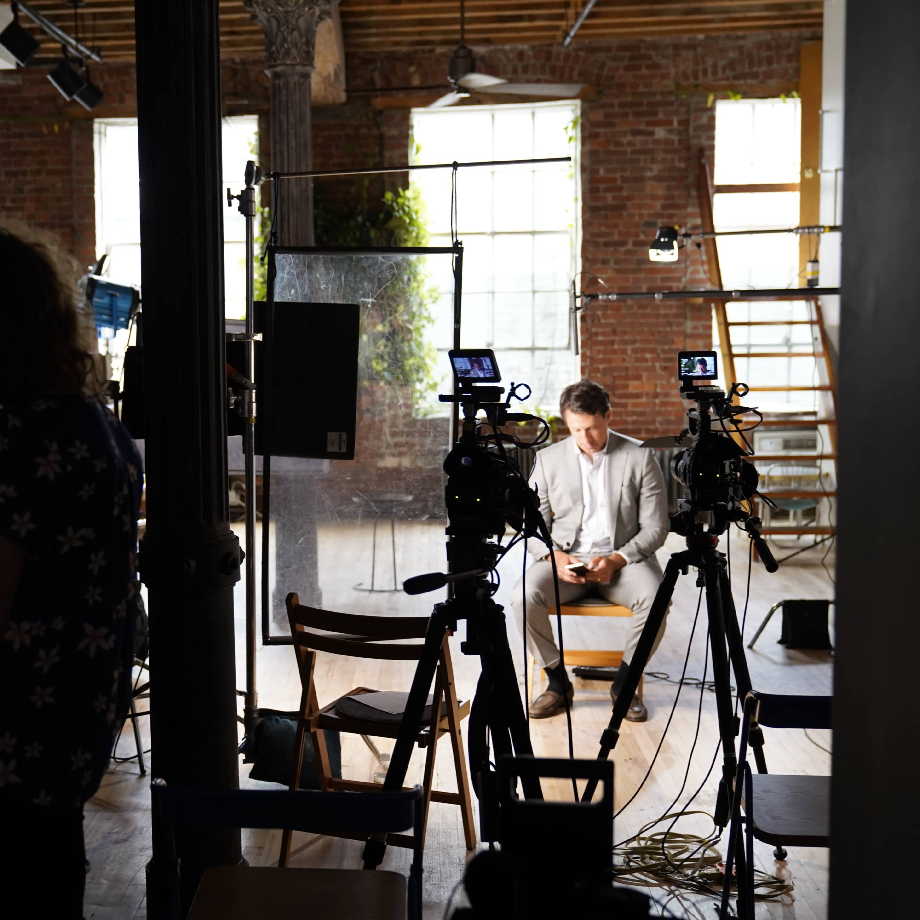 A man in a suit sits on a stool, looking at his phone, while cameras are set up around him in a studio with exposed brick walls.