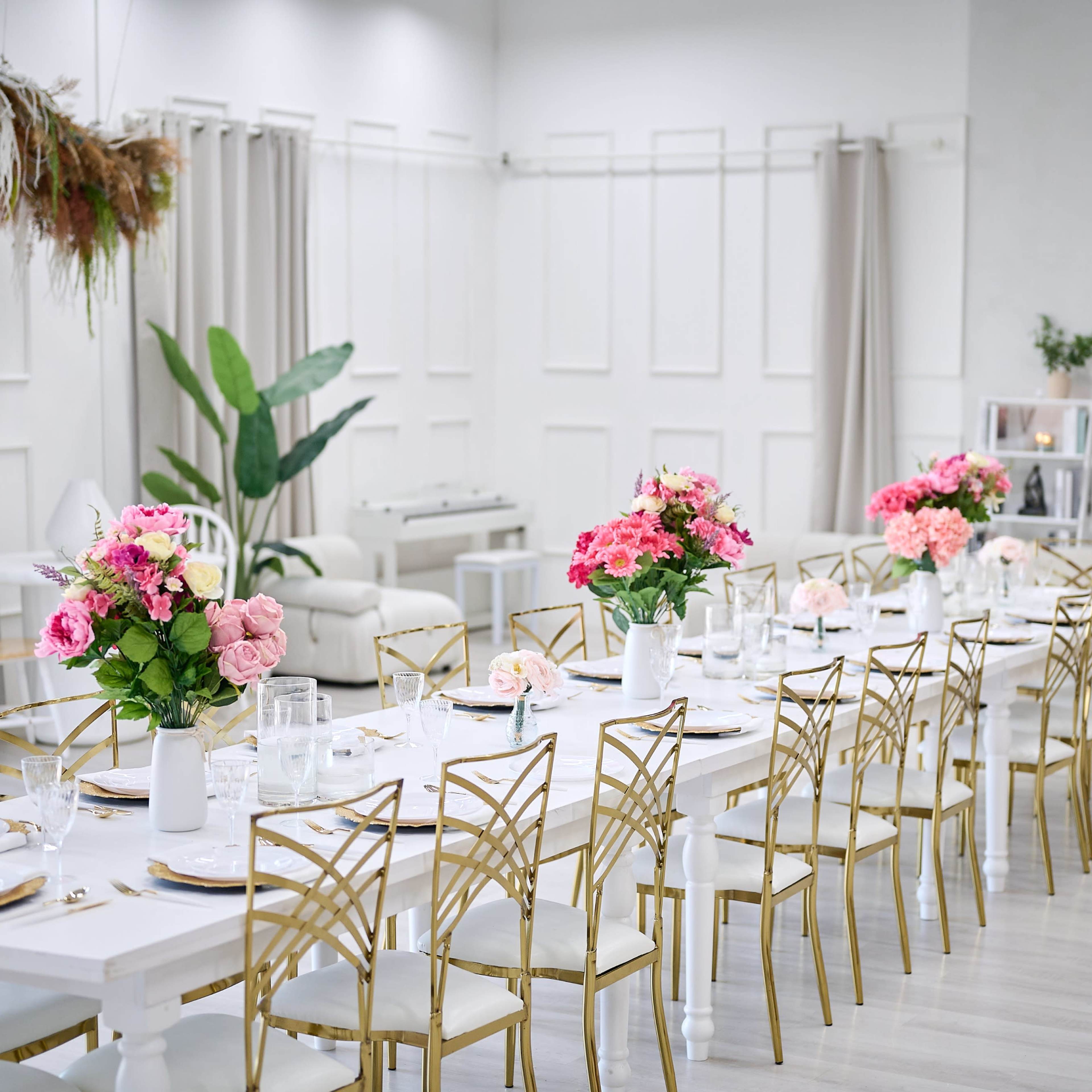 A long dining table adorned with pink flowers is set in a bright, modern space with neutral decor and greenery.